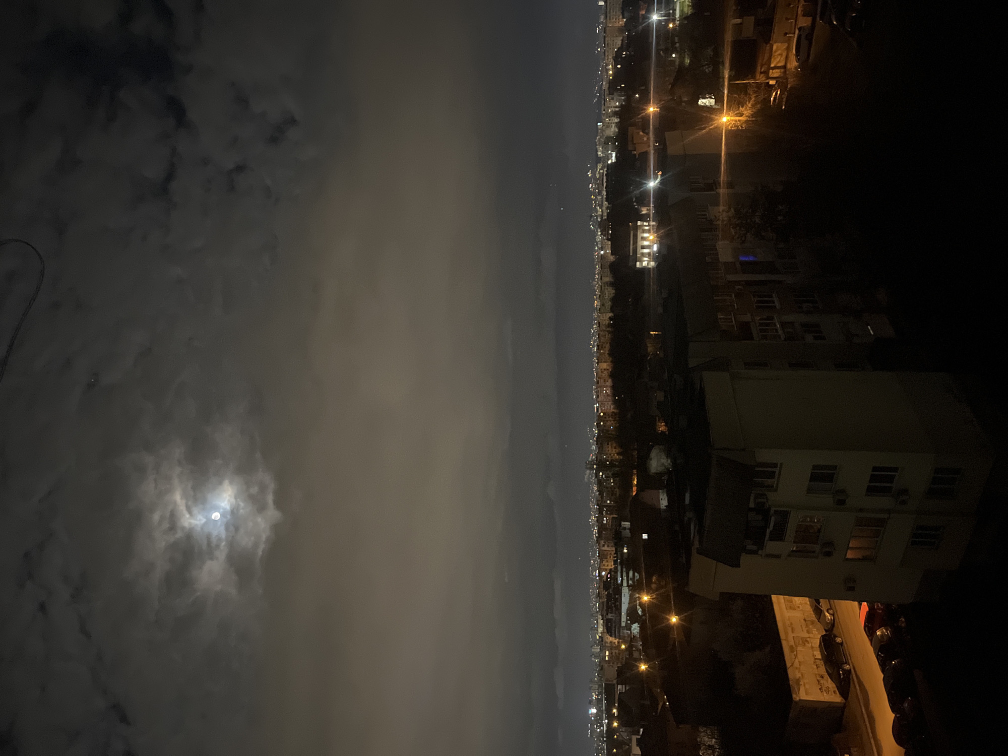 An eye-level long shot of the city of Sofia, Bulgaria, at night. The left side of the image depicts a cloudy sky. In the lower left corner, the moon is dimly visible through the clouds. The right side of the image shows a city skyline at night with illuminated streets and buildings. The photograph includes a view of buildings and cars in the foreground. Overall, the image depicts a cityscape under a cloudy night sky.