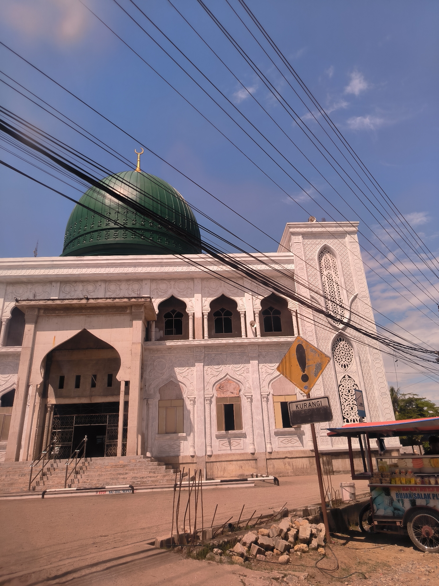 The image displays a large, white mosque featuring a prominent green dome crowned with a golden crescent moon finial, set against a vibrant blue sky with scattered clouds. Numerous black power lines cut across the upper part of the frame.
The mosque exhibits intricate architectural details, including ornate patterns carved into its white facade and a tall, slender minaret visible on the right. The main entrance is a grand arched structure with stairs and metal railings, appearing to be in an unfinished or renovated state with exposed concrete sections. Windows throughout the building also feature arched designs.
In the foreground, a dirt or gravel street runs alongside the mosque. On the right side, a yellow diamond-shaped sign with an exclamation mark is visible, mounted on a pole, with a black rectangular sign below it bearing the Indonesian word "KURANGI" (meaning "Reduce" or "Decrease"). Adjacent to the signs, a colorful, three-wheeled street vendor cart is partially visible, featuring text that appears to read "RUJAK SALAK PLU," suggesting it sells Rujak Salak (a fruit salad dish). Nearby, there is a pile of construction debris or bricks. Low, black and white striped barriers are positioned on the ground in front of the mosque steps; one barrier has the text "STOP SANDAL SEP," likely indicating a shoe-free zone before entering the mosque.
The scene is illuminated by bright daylight, suggesting a sunny day, likely late morning or early afternoon, in Keudelapang, Indonesia. There are no people visible, but the presence of the vendor cart and the construction materials hint at everyday activity around the mosque.