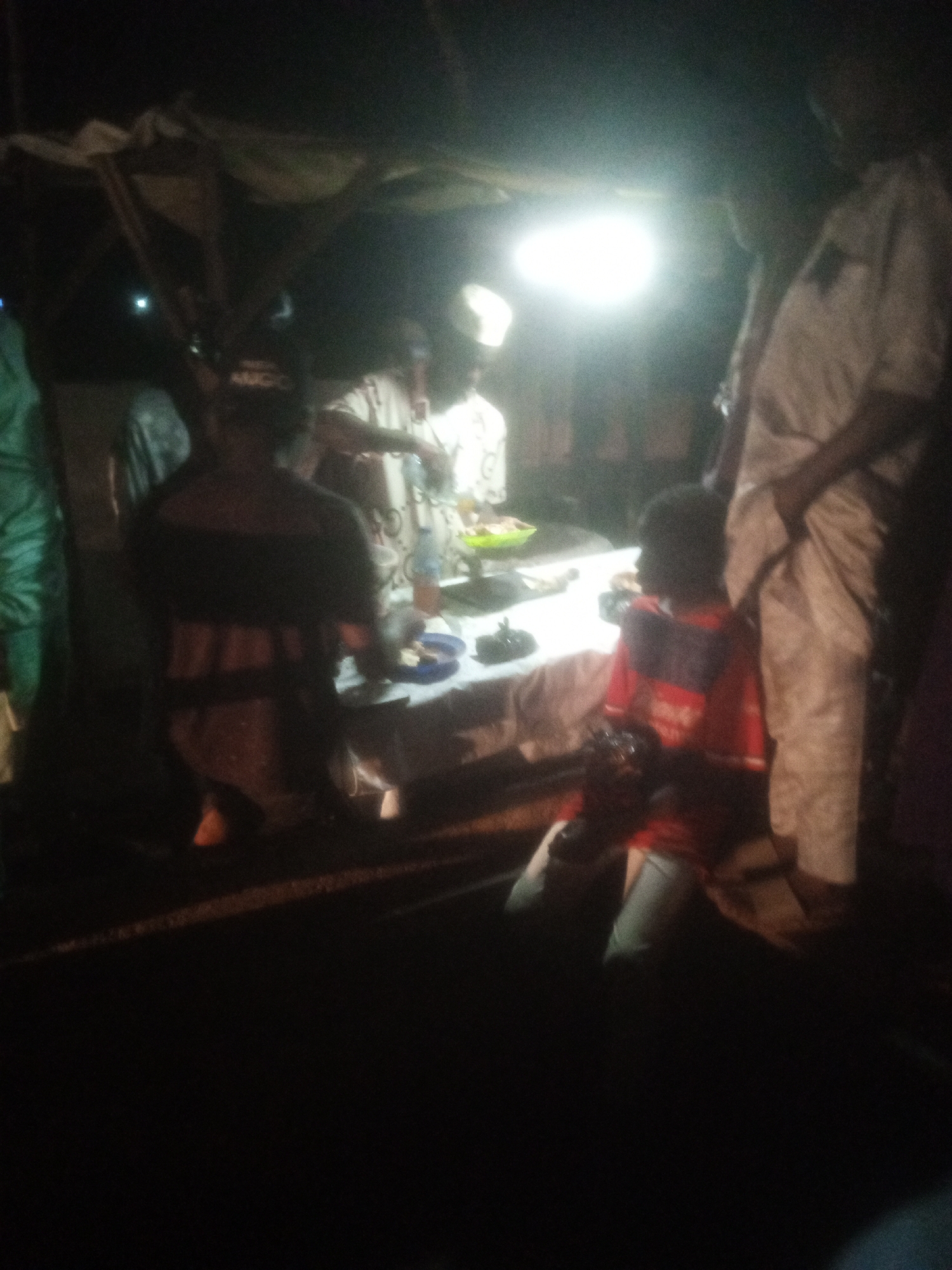 A dimly lit outdoor gathering is depicted in Tundun Wada West, Nigeria, under what appears to be a makeshift shelter, possibly a market stall. Several individuals are present, with a table set up in the center of the scene. The table is laden with plates of food, suggesting a communal meal or a food vendor. A bright light source illuminates the area, creating strong highlights and shadows.
In the foreground, a child in a red shirt is seated and looking towards the table. To the right of the child, an adult, partially obscured, stands beside the table. Behind the table, a group of people are gathered, some appearing to be interacting with the food or each other. The atmosphere suggests a social event or a busy marketplace at night. The background is dark, with hints of structures and vegetation, indicating an urban or semi-urban environment. The overall impression is one of a lively, albeit informally organized, nocturnal gathering.