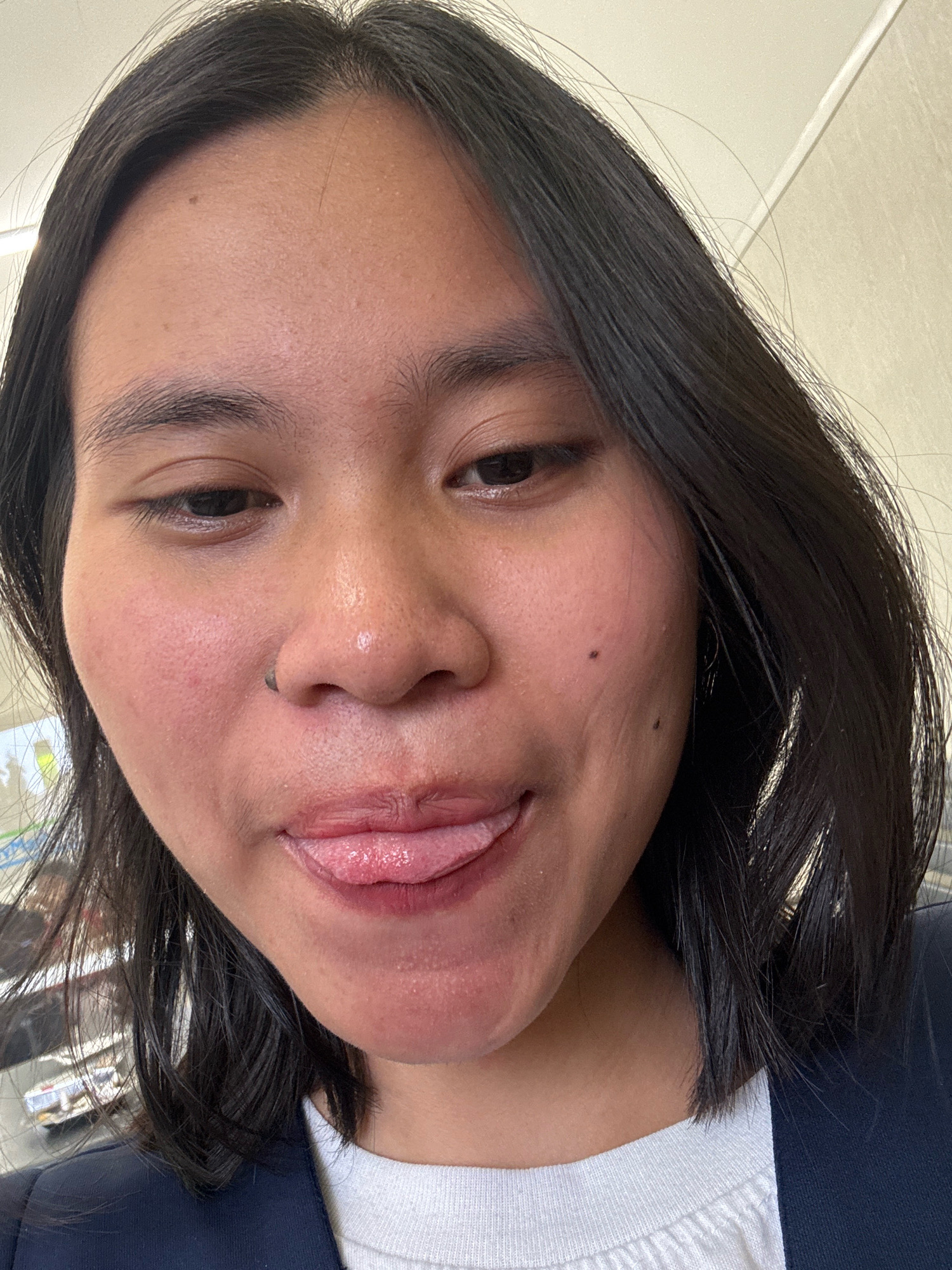 A close-up, front-facing selfie shows a person with dark, straight hair, and light skin. The person is wearing a dark navy blazer over a white t-shirt. Their eyes are open, and their tongue is sticking out. A small nose ring is visible. The background is a plain, light-colored wall. The image appears to have been taken indoors, possibly in an office or public building. The context of East Jakarta, Indonesia, is not visually evident from the image itself.