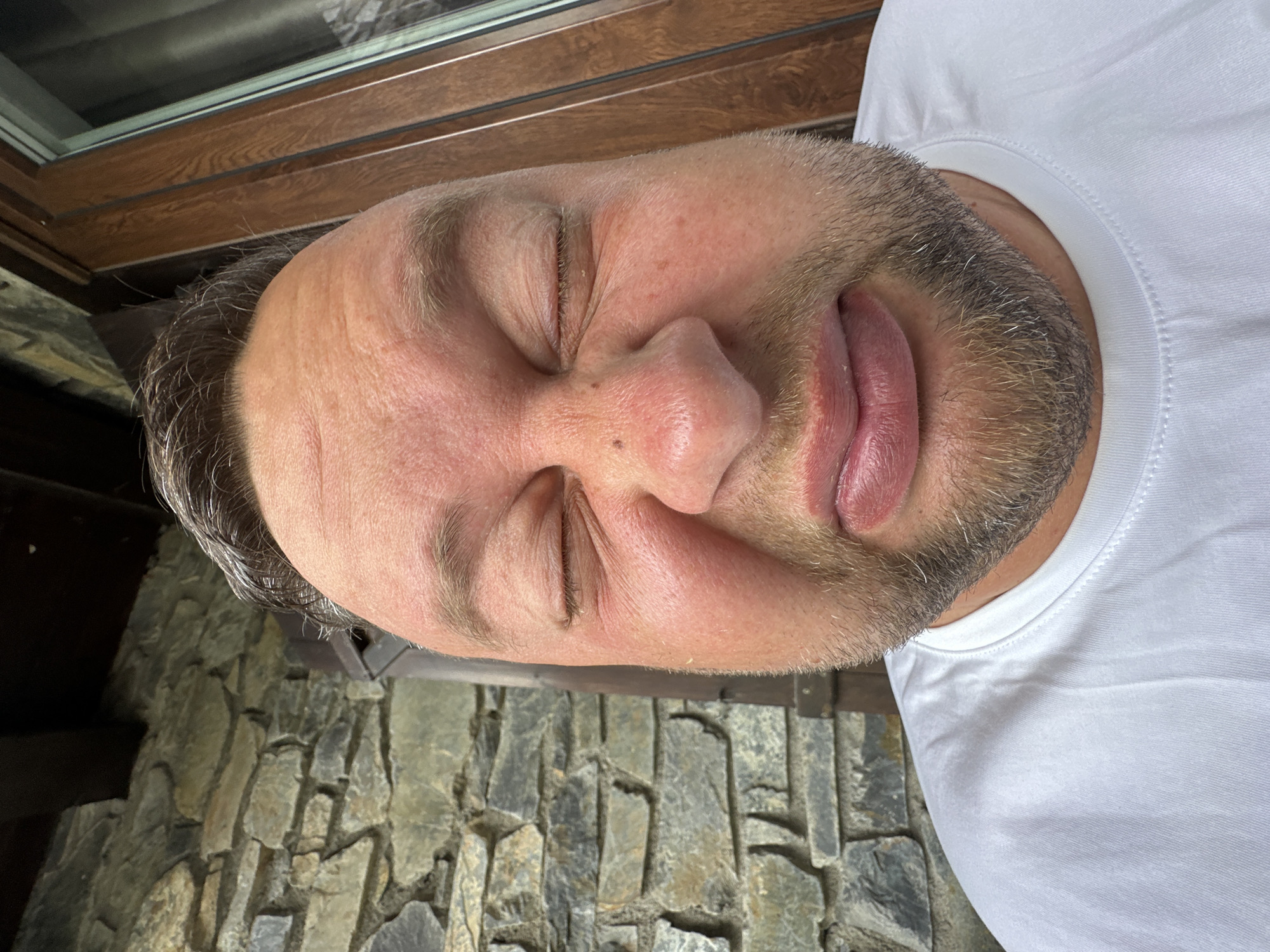 The image presents a close-up, tilted portrait of a man, likely taken indoors or under a covered area in Encamp, Andorra. The man, visible from his forehead to his upper chest, has fair skin, a short, light brown beard with hints of gray, and dark hair on his head. He is wearing a plain white crew-neck t-shirt.
His eyes are squeezed tightly shut, and his mouth is slightly open with his tongue partially extended. This exaggerated facial expression suggests a humorous or playful grimace, indicating feigned distaste, discomfort, or a comical reaction, rather than actual sleep.
The background reveals architectural elements: a dark brown wooden window frame is visible in the upper left, possibly a skylight or roof window. Below this, a rustic stone wall fills the background, composed of irregular, flat stones in various shades of gray, beige, and light brown. This type of stone masonry is consistent with mountain-style construction, fitting the given context of Andorra. The lighting appears soft and indirect. There is no visible text in the image.