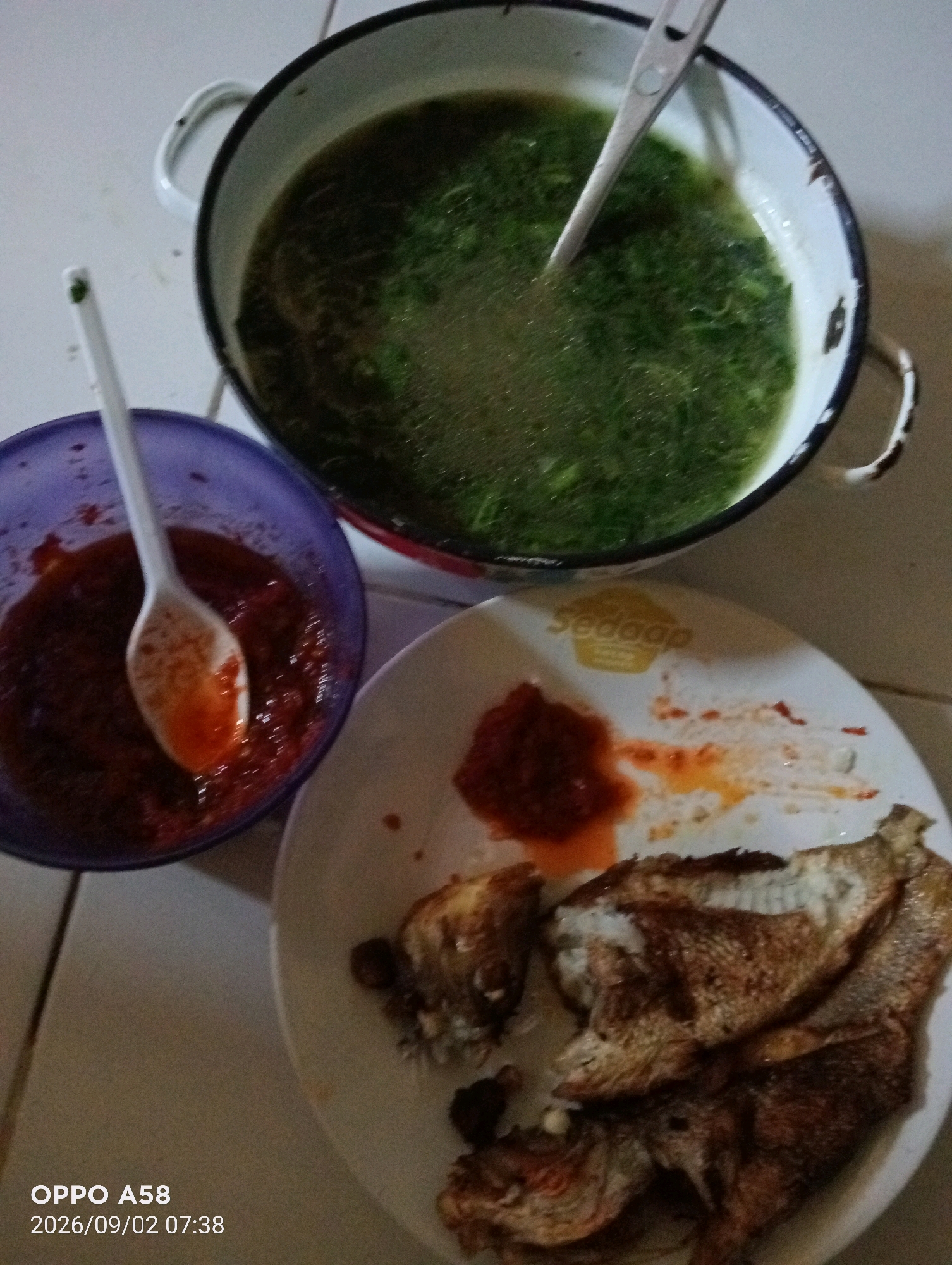 The image shows a meal laid out on a tiled surface, likely in the city of Pagerungan Kecil, Indonesia. The meal consists of a large pot of green vegetable soup, a small purple bowl filled with red chili paste and a white spoon, and a white plate with fried fish and more chili paste. The pot of soup is on the left side of the frame, and the bowl of chili paste is positioned below and to the left of it. The plate of fish is to the right of the chili paste bowl. The lighting suggests an indoor setting, possibly a home. The bottom left corner of the image displays a watermark reading "OPPO A58 2026/09/02 07:38".