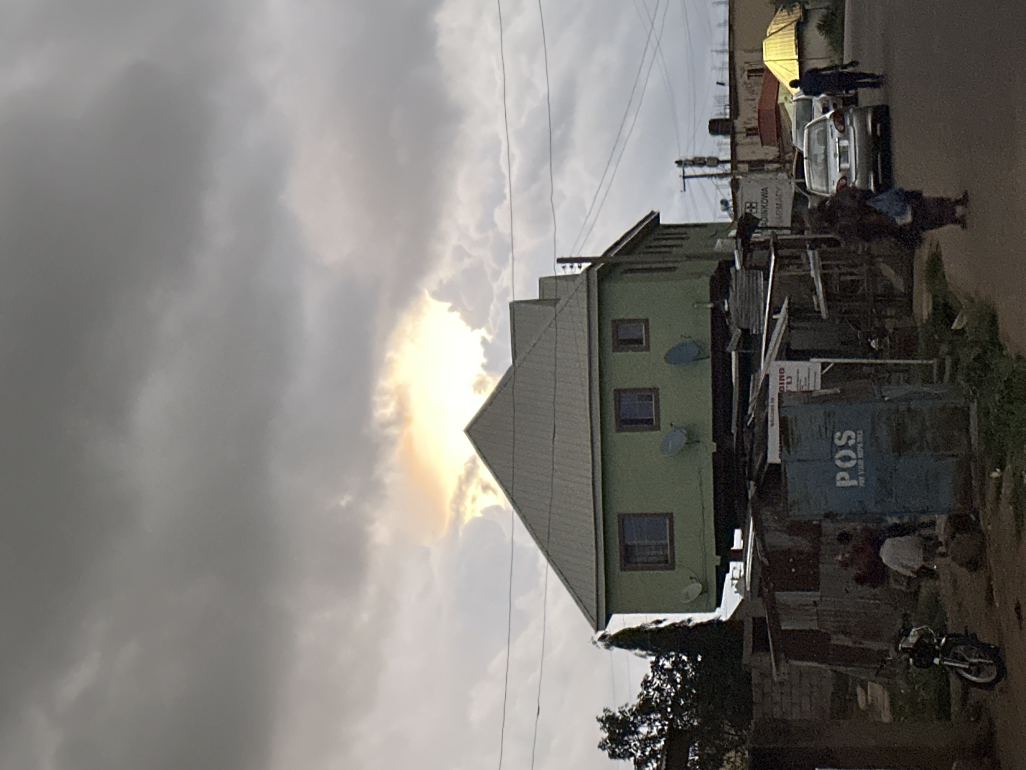 The image depicts an urban street scene under a heavily clouded sky, with a prominent patch of bright golden sunlight breaking through the clouds in the upper-middle. The precise location is unknown, but it represents a setting within the city of Unknown, Unknown.
In the center of the frame stands a two-story building with a light green exterior and a grey corrugated metal gabled roof. Four rectangular windows with brown frames are visible on its front facade, and two white satellite dishes are mounted below them. This green building appears to be elevated above a collection of smaller, more rudimentary structures constructed from corrugated metal and other materials, forming a base or surrounding complex.
On the left side of the foreground, a dark-colored motorcycle is parked on an unpaved surface next to a dark-leafed tree. Adjacent to this, in the mid-ground, is a blue, rectangular structure with "POS" and "PAY YOUR NEPA BILL" painted in white, indicating a point of sale or service. Several individuals are visible near and behind this structure, one wearing a white top.
To the right, an asphalt road occupies the middle and right foreground. A silver sedan is parked or moving slowly on the road, facing away from the camera. Another vehicle is partially obscured behind it. Two individuals are walking on the road: one in dark clothing on the right, moving away from the viewer, and another in dark clothing on the left, moving towards the viewer, carrying a blue item.
Utility poles and numerous overhead power lines crisscross the scene, particularly on the right. A white signboard featuring text including "ADINKOWIA PHARMACY" is affixed to a pole near the silver car. In the background on the right, a building with a distinctive yellow, possibly metal or tarp, roof is visible, along with other unpainted or faded structures. The ground in the foreground is uneven, with some sparse vegetation and a visible ditch or gutter.