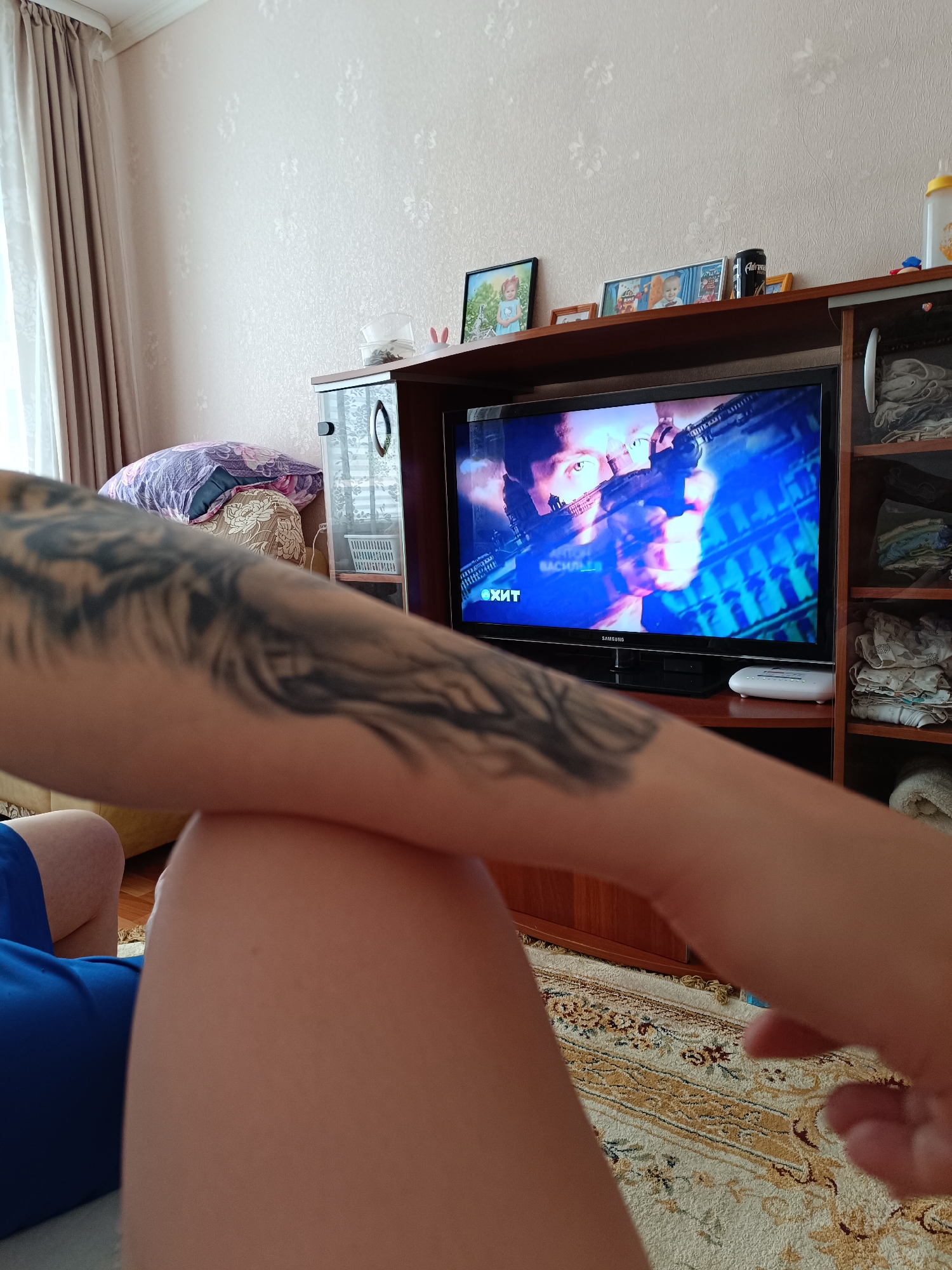 A person with a tattoo on their arm is seated indoors in Tobolsk, Russia, watching a Samsung television. The TV is displaying a scene from an action movie with a man holding a weapon. The TV is positioned on a wooden cabinet with photos and objects on top. A rug is visible on the floor. Curtains partially cover a window in the background.