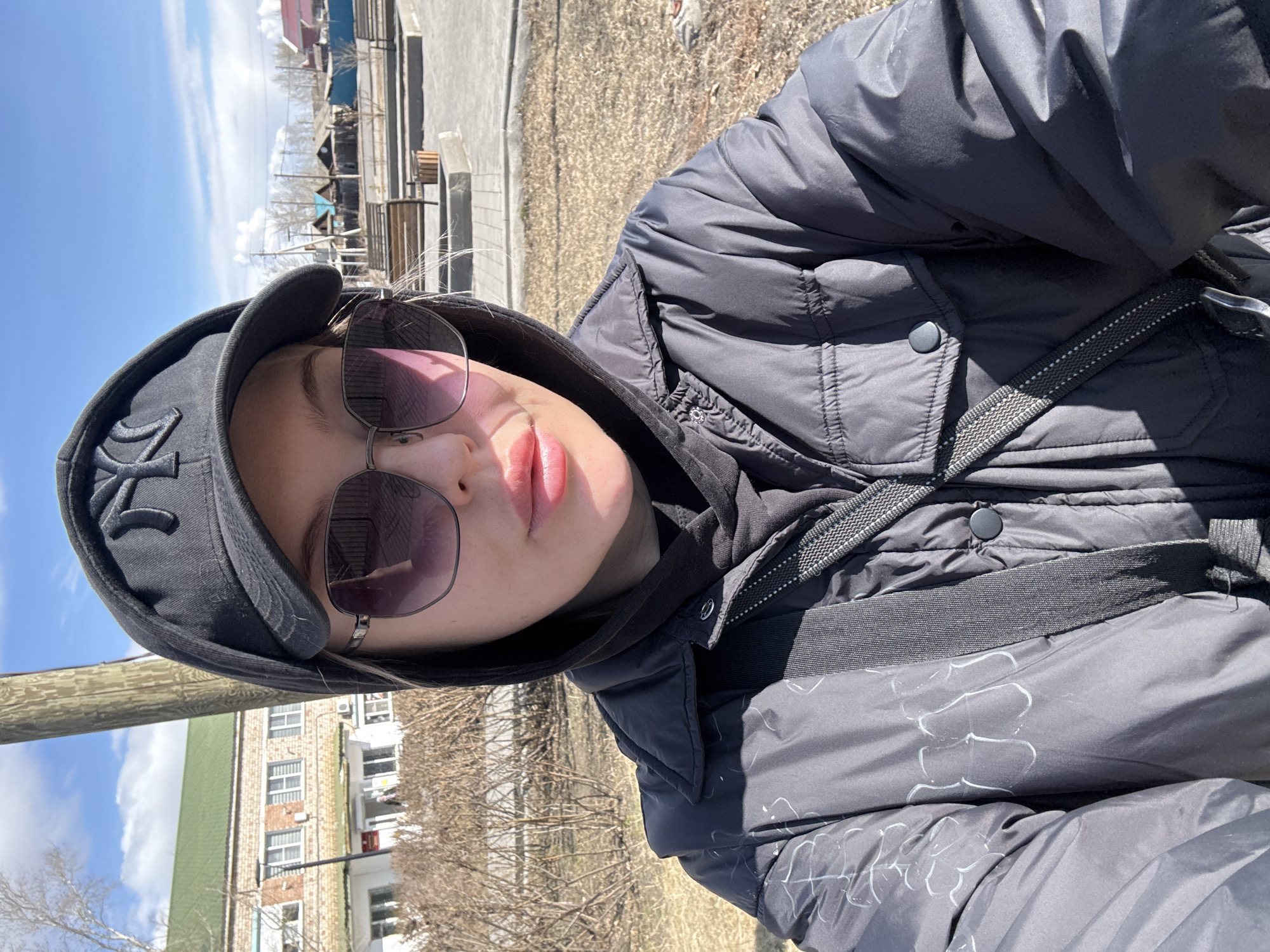A person wearing a black jacket, a black hoodie, sunglasses, and a black baseball cap is facing the camera. The person is outdoors on a sunny day in Borzya, Russia. In the background, a brick building with windows is visible, as well as a wooden structure with a blue roof. The person is standing on a patch of grass or dirt, and a wooden post is visible to the left of the person.