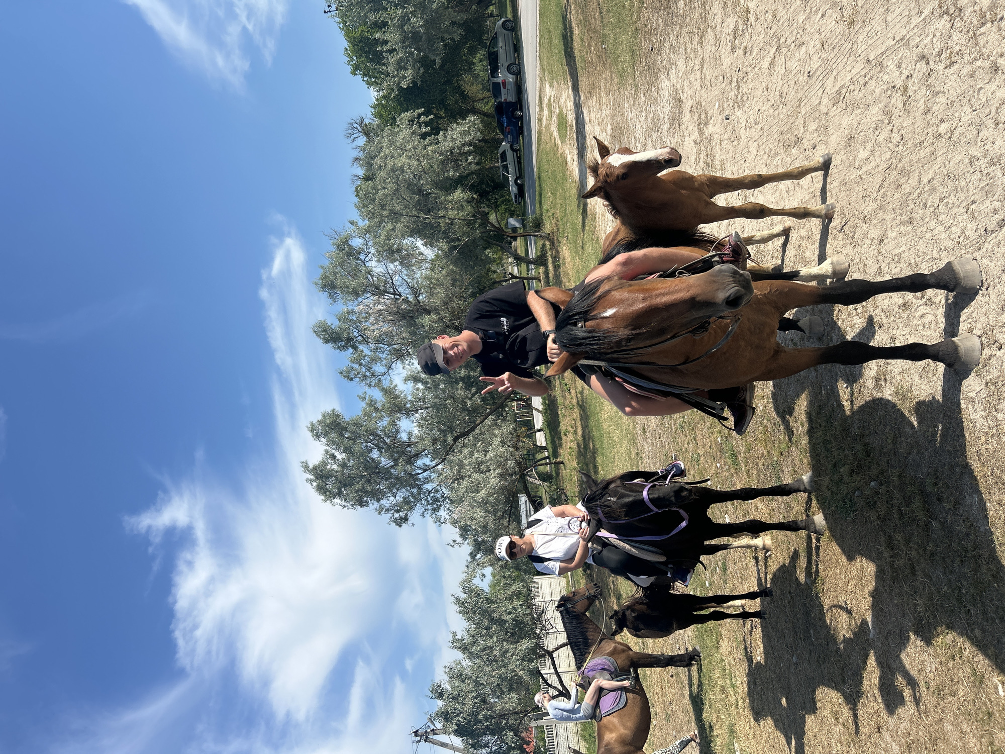 In Мысовское сельское поселение, Ukraine, under a bright blue sky with scattered white clouds, a group of people are enjoying horseback riding. In the foreground, a man wearing a black t-shirt and a black cap rides a brown horse and makes a peace sign with his hand. To his right, a brown foal stands by another horse. Behind the man, a woman in a white t-shirt and a white cap is on a black horse. To the left, a person on a brown horse, wearing a light blue long-sleeved shirt and grey shorts, rides with a black foal beside them. In the background, several cars are parked along a road, and trees line the area. The ground is a mix of dry grass and dirt paths, with shadows cast by the horses and riders.