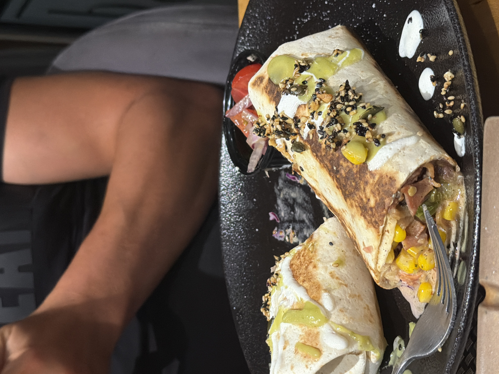 Two burritos are presented on a black plate. The burritos are filled with corn, meat, and vegetables, and topped with a creamy white sauce and a mixture of seeds. A fork is resting on the plate, next to one of the burritos. In the background, a person is partially visible, wearing a dark t-shirt. A small dish of chopped tomatoes and red onions is also on the plate. The scene appears to be in an indoor setting, possibly a restaurant. There is no information available to determine the specific location in Plovdiv, Bulgaria.
