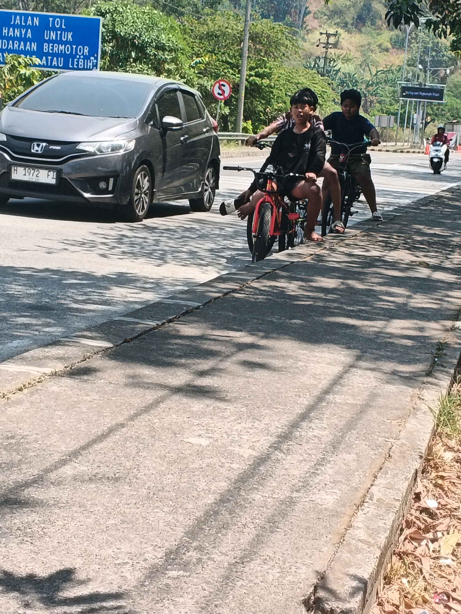 On a sunny day in Kalirejo, Indonesia, two young males are riding on separate small motorcycles. The person in front, on a red motorcycle, is wearing a black long-sleeved shirt and shorts. The second rider, on a darker motorcycle, is wearing a navy blue t-shirt and shorts. Both are looking forward, and the second rider appears to be slightly behind and to the right of the first. They are riding on a paved road, likely a highway, with a white pedestrian crossing marked on the asphalt. A dark gray Honda hatchback car is parked on the left side of the road, and a blue highway sign is visible in the background. The environment is characterized by lush green trees and vegetation on a hilly terrain. The sun casts distinct shadows on the road, indicating the time of day is likely midday.