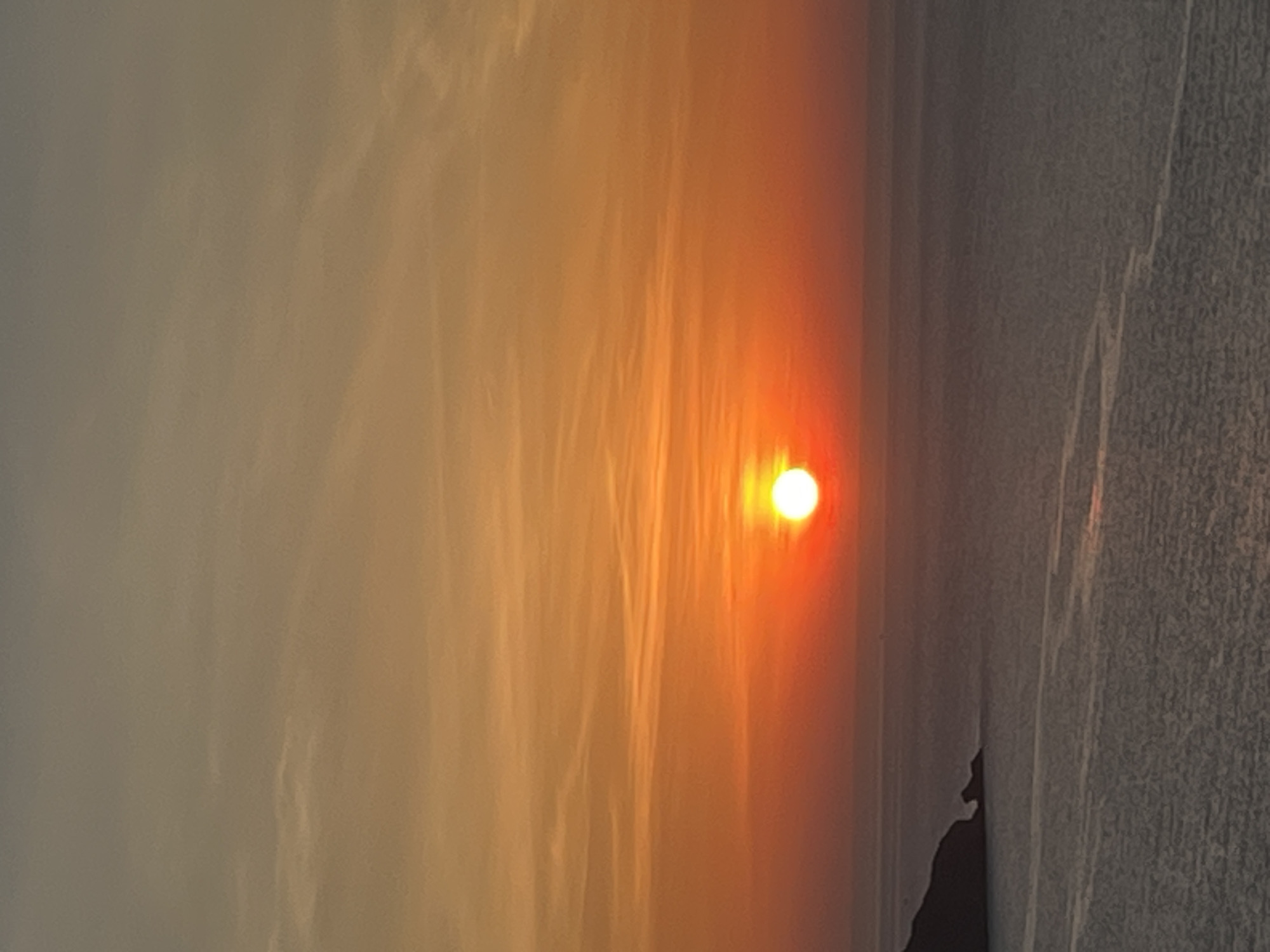 This is a vertical, eye-level, medium shot of a sunset over the ocean. The sun, a bright white orb with an orange halo, is positioned slightly left of center in the frame, halfway up the image. It is casting a warm orange glow across the sky, which transitions from a pale yellow at the horizon to a muted gray at the top. Wispy clouds streak horizontally across the sky.
In the lower left corner of the image, a dark, rocky peninsula juts out into the calm, gray ocean. The water reflects the colors of the sunset, with hints of orange and pink visible in the subtle ripples.
The scene appears to be taken from a high vantage point, possibly a cliff or a building overlooking the coastline. The location is likely along the coast of Đà Nẵng, Vietnam, given the presence of the ocean and the general atmospheric conditions associated with sunsets in the region. There are no people or any specific objects or landmarks visible that would definitively pinpoint the exact location within Đà Nẵng. The overall atmosphere is serene and tranquil.