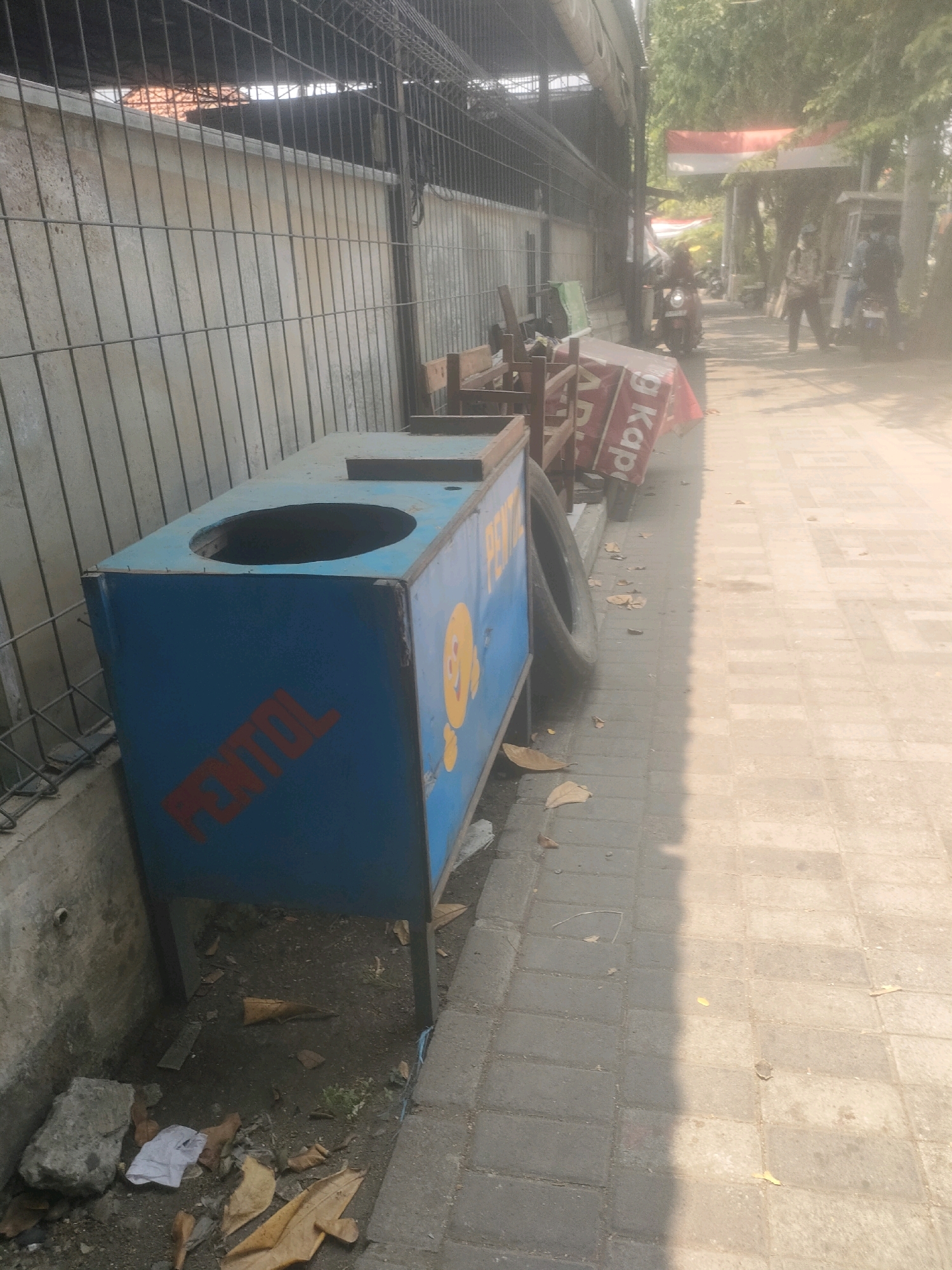 The image depicts an outdoor scene on a sunny day, likely in Surabaya, Indonesia, characterized by its urban setting with paved walkways and a fence. In the foreground, a blue rectangular bin with a circular opening on top is prominently displayed. The bin has red lettering that reads "FENTOL" on its side, along with a yellow cartoon character. Adjacent to the bin is a discarded tire. The ground around the bin is littered with dry leaves and debris, suggesting a lack of recent cleaning.
Behind the bin, a wire fence runs along a concrete wall, partially obscuring a building or structure covered by a dark corrugated roof. Further down the paved walkway, there are several objects, including what appears to be a wooden stand and a red banner with white lettering, partly visible and leaning against a structure.
In the background, the scene suggests a public area with trees providing shade. Several individuals are visible, some on motorcycles and others walking. One person is riding a red motorcycle, while others appear to be standing or walking along the path. A red and white Indonesian flag is hung across a pathway in the distance, indicating a possible national holiday or event. The overall atmosphere is one of everyday activity in an outdoor urban environment.