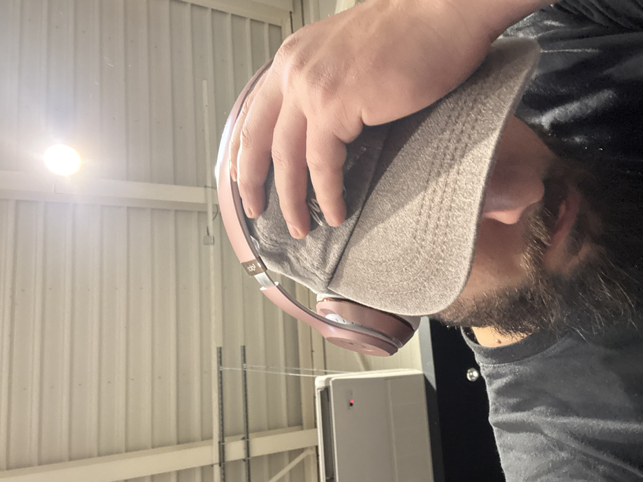 The image depicts a male individual in an indoor setting in Strasbourg, France. The subject is shown from the nose area down to the chest, wearing a dark t-shirt, a grey baseball cap pulled low, and rose gold over-ear headphones positioned over the cap. A dark beard is visible on his face. His right hand is placed on the side of the cap. The background features a white corrugated metal ceiling with visible support structures and suspended wires. A bright, unshielded overhead light source is present in the upper right. In the upper left background, a white wall-mounted unit, likely an air conditioning or ventilation system, with a small red indicator light, is visible. The individual's head is oriented downwards, obscuring the eyes.