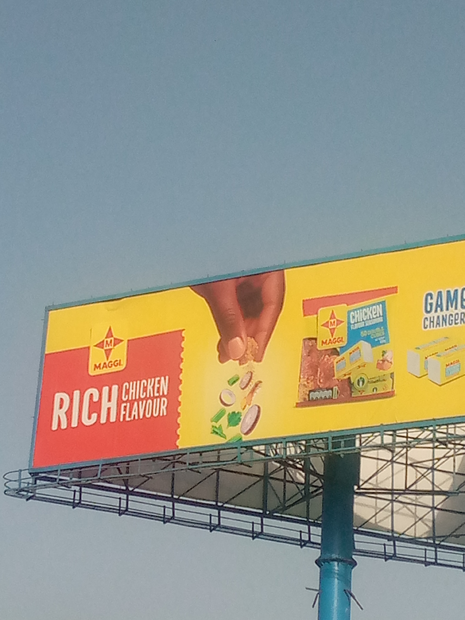 A billboard advertising Maggi chicken flavor seasoning stands prominently against a light blue sky. The billboard features the Maggi logo, text proclaiming "RICH CHICKEN FLAVOUR", an image of a hand sprinkling seasoning, and a depiction of Maggi seasoning products. The structure supporting the billboard is composed of metal beams and a blue pole. This image was taken in Bukuru, Nigeria.