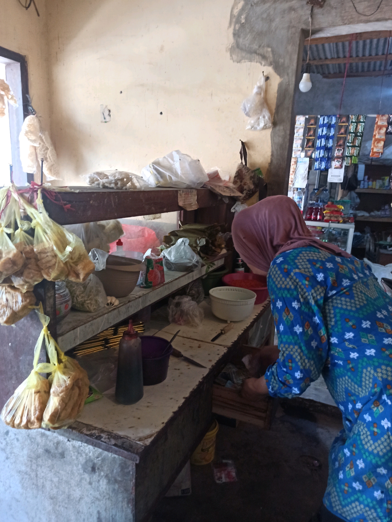 The image depicts an indoor market stall in Tlogosari, Indonesia. A woman wearing a pink headscarf and a blue patterned tunic is standing behind a counter, seemingly preparing food. The stall is filled with various items, including bags of food products hanging from the ceiling, bowls, bottles of condiments, and packaged goods displayed on shelves in the background. The environment appears somewhat cluttered but functional, suggesting a typical small business operation. The lighting is a mix of natural light from an unseen window and artificial light from a hanging bulb.