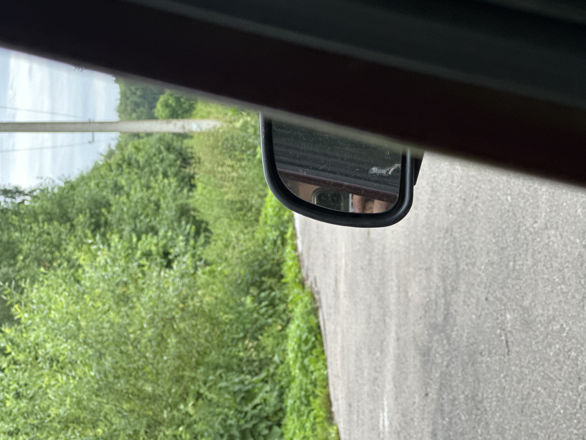 A side-view mirror of a car reflects a person holding a phone. The car appears to be on a paved road with a tree line on the left side and a utility pole in the background. The overall scene suggests a view from inside a vehicle driving through a rural or suburban area in Vostets SNT, Russia.