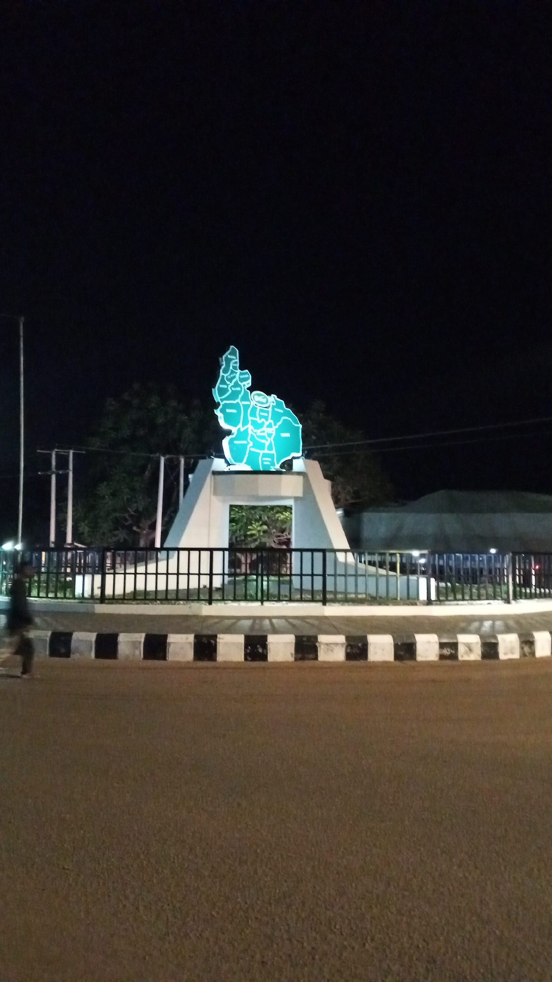 A nighttime shot captures a roundabout in Bukuru, Nigeria. The roundabout's central feature is a white concrete structure supporting a turquoise map of Plateau State outlined in lights. A black metal fence encircles the base of the structure, behind which is a planting of greenery. The curb is painted in alternating black and white blocks. A person walks by on the left. The road is asphalt. Trees are visible behind the roundabout structure. The sky is black and lights up the structure.