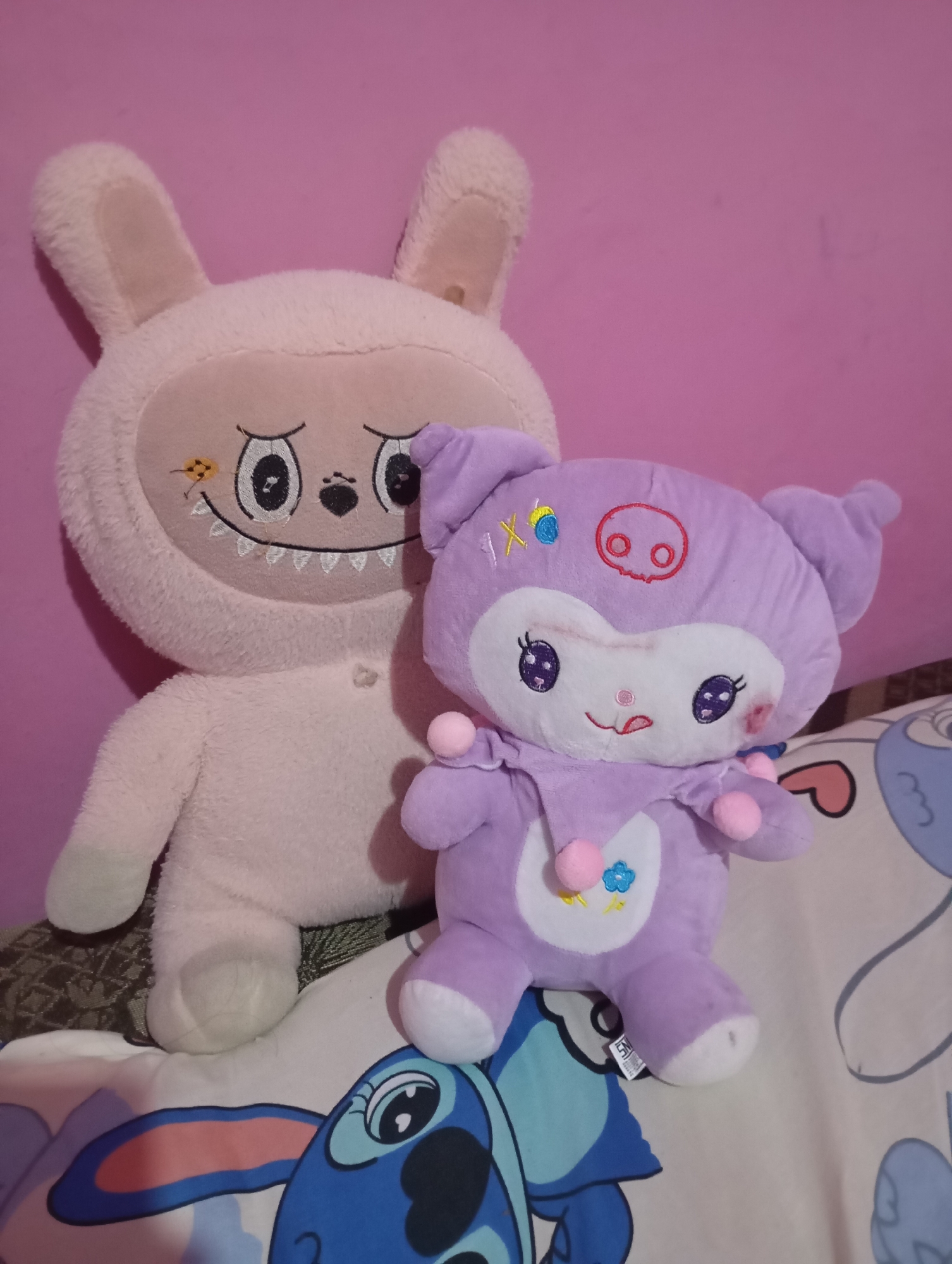 Two stuffed toys are positioned on a bed against a pink wall. The larger stuffed toy on the left is pink with rabbit ears and has a detailed embroidered face featuring wide eyes, a nose, and a wide grin with sharp teeth. It is wearing a fuzzy jumpsuit. To the right of the pink toy is a smaller, lavender-colored stuffed toy with cat-like ears and a pig-like nose. This toy has large, expressive eyes, small pink pom-poms around its neck, and a flower detail on its chest. The bedspread beneath the toys features a pattern of cartoon characters, including Stitch from Disney's Lilo & Stitch. The scene is indoors, and the lighting suggests it is daytime. The location in Leman Benawi, Indonesia, is not visually discernible from the image itself.
