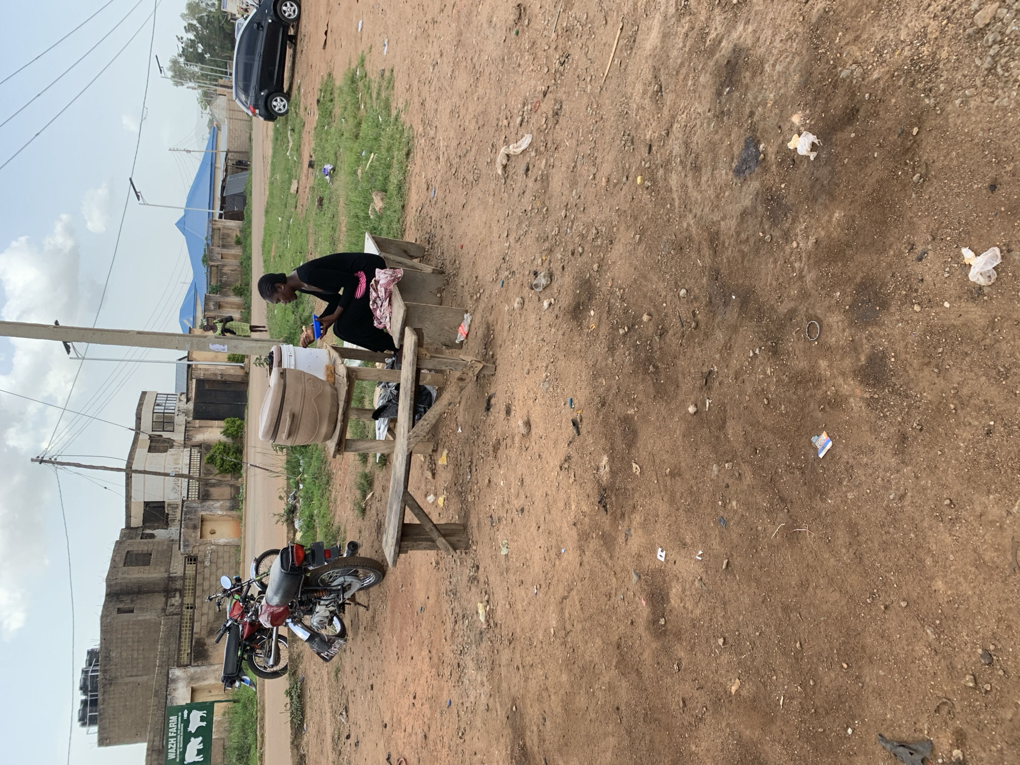 The image depicts an outdoor scene in Barakin Lerawi, Nigeria. In the foreground, a young woman wearing a black dress sits on a concrete bench, holding a blue bowl and a utensil, appearing to eat. A wooden table with a large plastic container is positioned to her left. Behind the woman, two motorcycles are parked. To the far left, a sign reads "WAZH FARM" with images of livestock. In the background, buildings with brick exteriors and some unfinished construction are visible. A black car is parked on the right side of the street. The ground is primarily dirt with scattered debris. The sky is overcast.