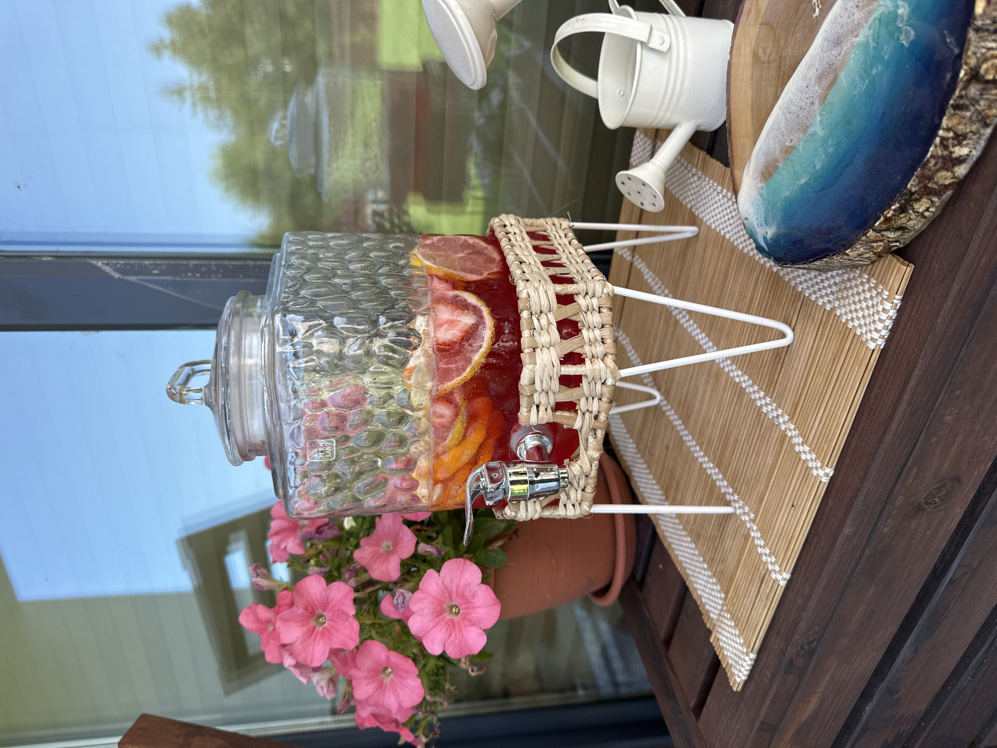 This is a close-up, eye-level outdoor shot of a glass beverage dispenser filled with a red liquid and fruit slices, likely on a wooden table. The dispenser has a metal spigot and a woven basket base supported by white legs. Behind it, a pot of bright pink flowers adds a splash of color. To the right, a small white watering can sits on the table. In the foreground, a piece of wood with a resin art depicting a wave is visible. The background shows a reflection of trees and sky in a large window or glass door. The overall scene suggests a relaxed, possibly summery, outdoor setting. The location is not specified in the image or context.