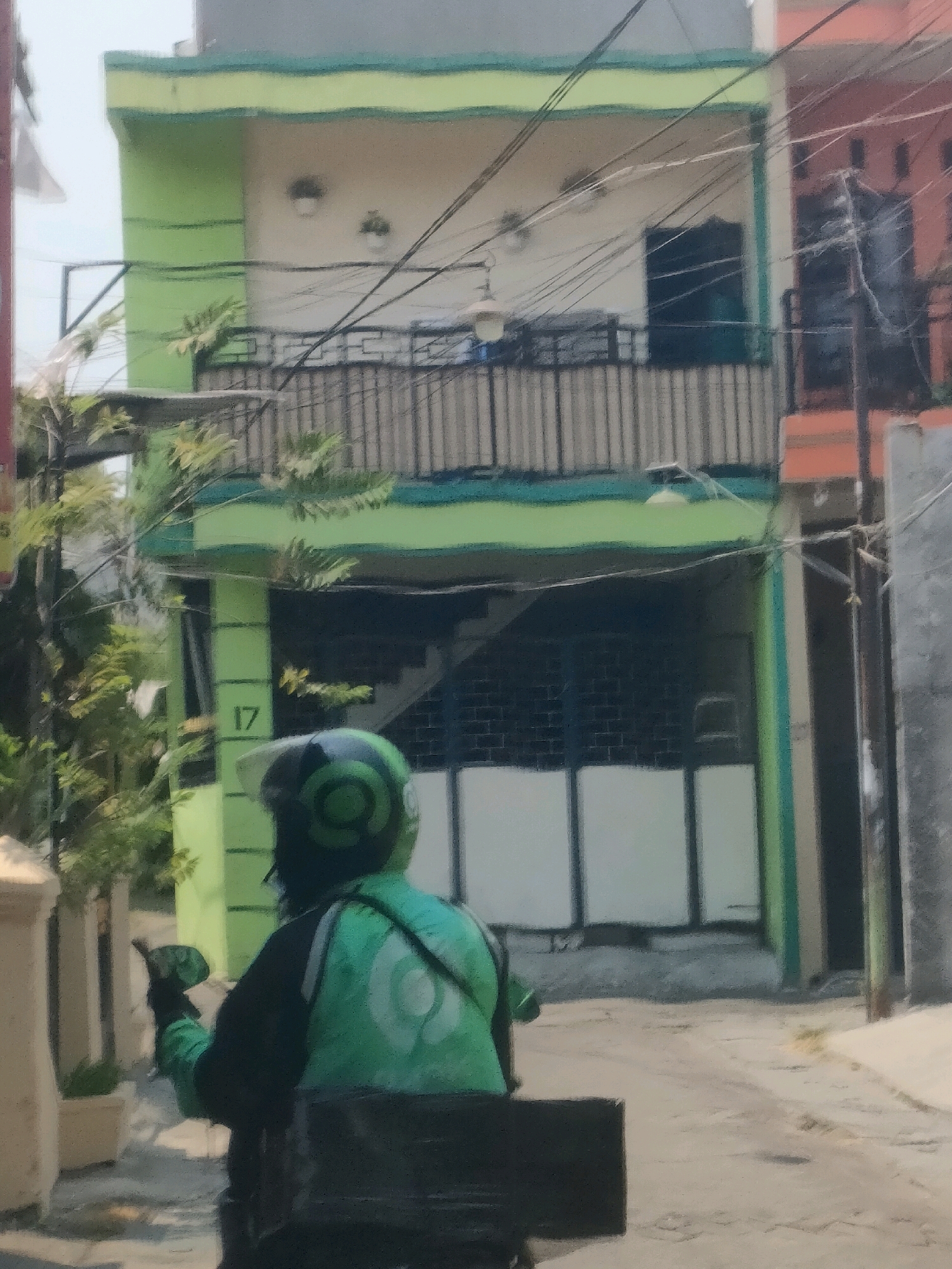 The image shows a person wearing a green and black Gojek uniform, including a helmet with a green Gojek logo, riding a motorcycle. The person is viewed from behind and appears to be in a street. Behind the motorcycle rider, there is a light green building with a balcony and what appears to be a garage door. The building has the number "17" visible on its exterior. The street appears to be paved. The surrounding environment suggests an urban setting, possibly in Surabaya, Indonesia, given the Gojek branding which is prevalent in Southeast Asia. There are also power lines visible overhead, and adjacent buildings. The lighting suggests it is daytime, likely with overcast skies.