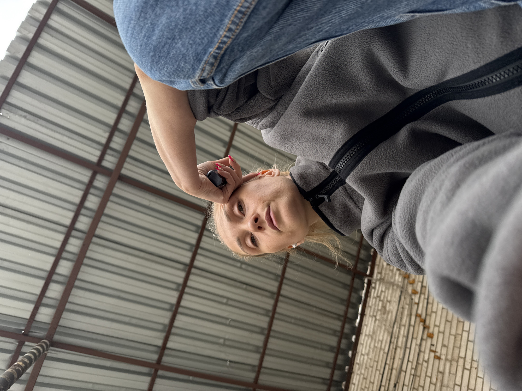 The photograph presents an upward-looking perspective. A person, presumably the photographer, is looking down at the camera. The person is wearing blue jeans and a gray fleece jacket with a black zipper. The person is holding a small black object near their forehead.
The background consists of a metal roof with brown support beams. The lower right corner of the image shows a portion of a brick wall.
The location is Chaikovsky, Russia. No further specifics can be derived about the exact location.
