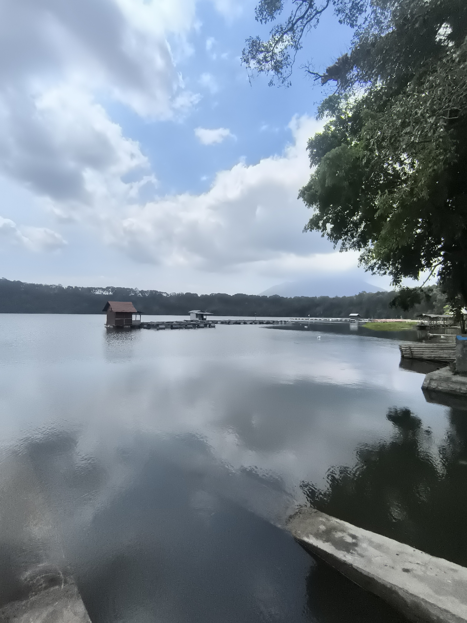 This is a medium shot, exterior, vertical, eye-level, day time photograph of a serene lake with a small wooden hut on stilts in Tegalrandu, Indonesia. The lake's surface is calm and reflects the overcast sky and surrounding trees. In the foreground, there is a concrete structure entering the water from the right side of the frame. A long wooden pier extends from the hut into the lake. Dense green trees line the far bank of the lake, and a mountain range is visible in the background, partially obscured by mist. The sky is filled with scattered clouds, some dark and ominous, others bright and white. The overall atmosphere is tranquil and natural. There are no people visible in the image.