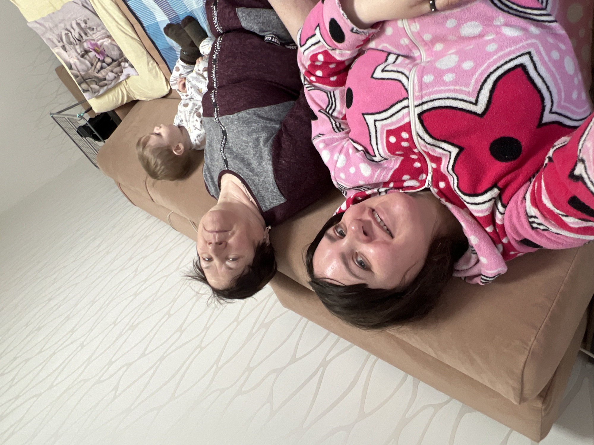The image shows a family scene indoors. There are three people visible: two adults and a young child.
The first adult, possibly a grandmother, is wearing a maroon and gray outfit. The second adult, potentially a mother, is in a pink and red patterned pajama top. The child is wearing a white patterned onesie and brown socks. All three are lying on a brown couch.
The location appears to be a home, possibly in the city of Perm, Russia, based on the information provided in the prompt. The background includes a wall with a light-colored, patterned wallpaper. In the upper left corner, there is a bed with multiple pillows and blankets. A clothes drying rack is also visible in the background.