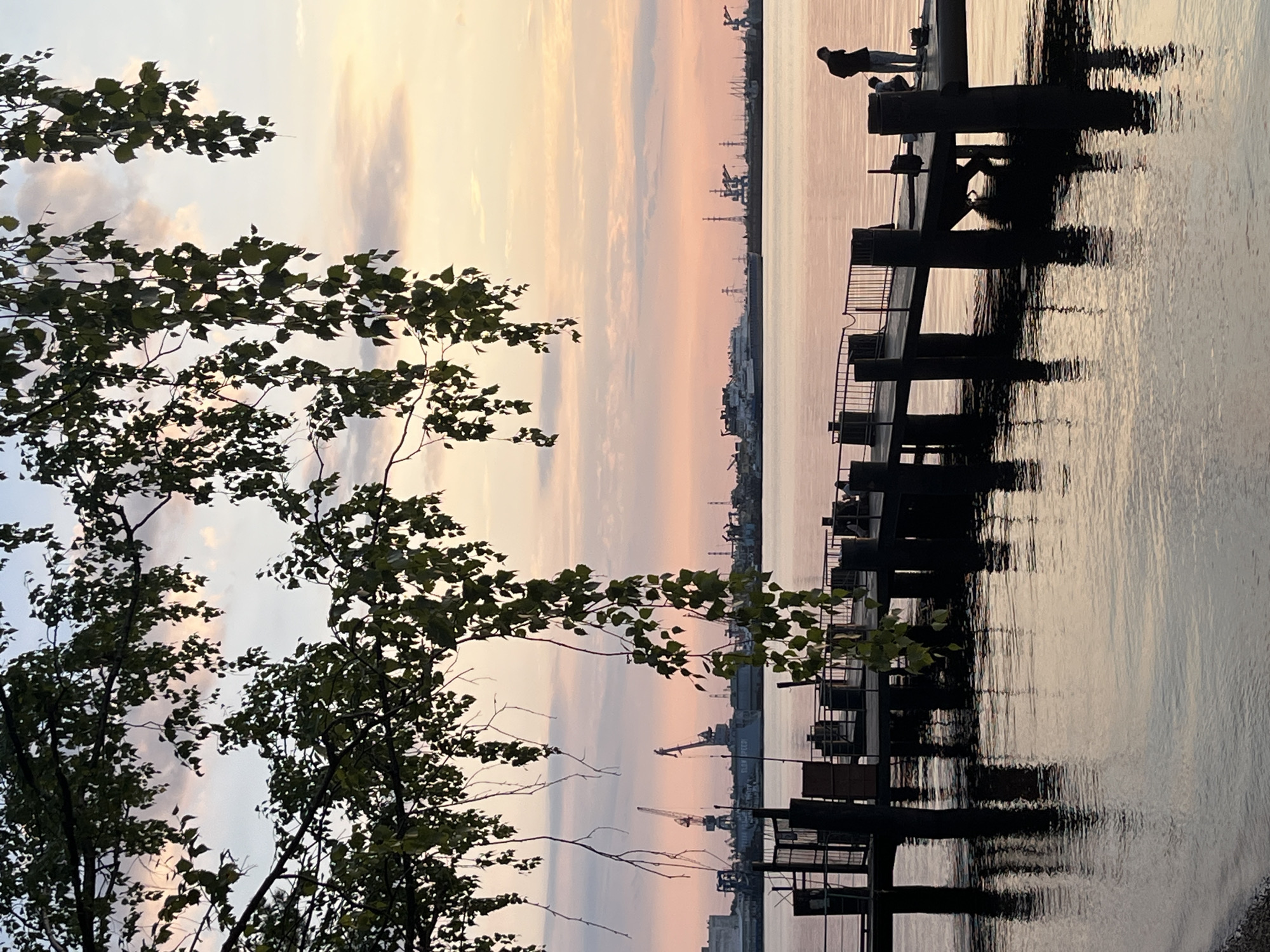 The image depicts a sunset scene at a pier on the Kama River in Chaikovsky, Russia. In the foreground, a dark wooden pier extends into the water, supported by thick pilings. The pier has a railing along its side, and a few dark structures are visible on it. Two individuals are present on the pier; one stands with their back to the camera, while another person is crouched down. Overhanging branches with green leaves frame the top of the image, casting shadows on the sky. The sky is a soft blend of pink, orange, and pale blue, with scattered clouds. In the distance, across the river, a hazy industrial skyline with cranes and ships is visible. The water reflects the soft colors of the sky and the dark pier.