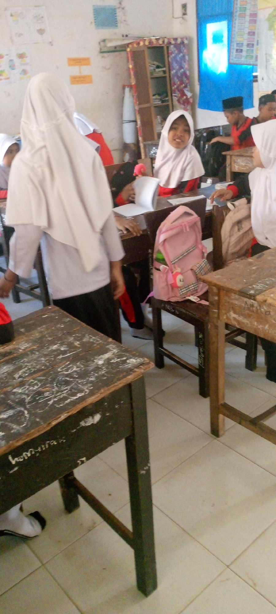The image depicts a classroom setting in Baratpayaitek, Indonesia, with several students, predominantly young girls wearing hijabs, present. In the foreground, a dark wooden table with visible wear and tear, including graffiti, is positioned on a tiled floor. To the right, another wooden table and chairs are visible. A pink backpack with a checkered pattern and a small charm is placed on a chair. In the mid-ground, several students are seated at tables. One student in the center is looking directly at the camera and smiling, wearing a light-colored hijab and a red garment. Other students are partially visible, some interacting with books and papers on their desks. The background reveals a whiteboard, shelves with various items, and posters on the walls, suggesting an educational environment. The lighting appears to be natural, coming from a window or overhead sources, casting soft shadows. The overall impression is one of a typical Indonesian school classroom.