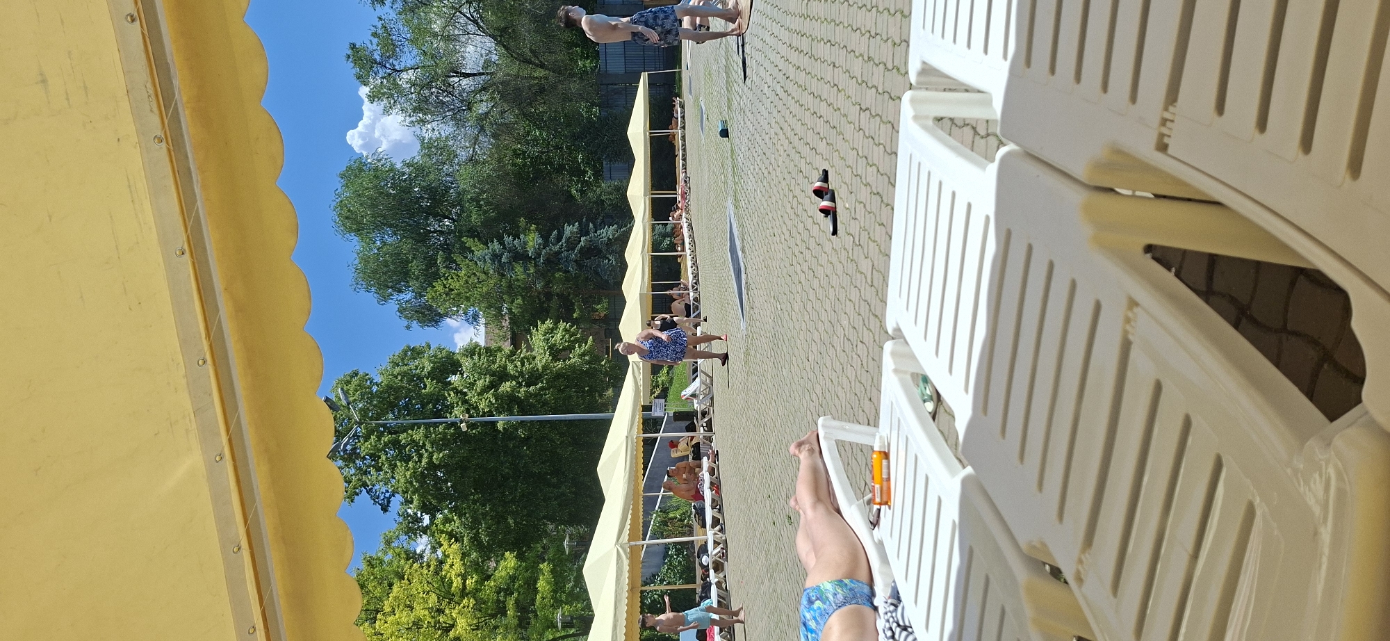 The image depicts a sunbathing area at an outdoor aquatic facility, likely a water park or a public pool, in Almaty, Kazakhstan. In the foreground, two white plastic lounge chairs are unoccupied and arranged side-by-side. A person's legs are visible on the chair to the left, wearing blue and white patterned swimming trunks. An orange bottle of sunscreen and a pair of sunglasses rest on this chair.
In the middle ground, a paved walkway with interlocking hexagonal stones stretches towards a row of identical white tents supported by metal poles, providing shade. Several people are seated or lying on lounge chairs beneath these tents. A woman in a blue and white patterned dress walks across the paved area. To her left, a young boy in blue swimming trunks stands near the edge of the shaded area. Further back, a young man in patterned swimming trunks stands on the right side of the frame. Scattered across the paved area are some casual flip-flops.
The background features lush green trees and a bright blue sky with scattered white clouds. A lamppost is visible between the trees. The overall impression is that of a warm, sunny day at a recreational venue.