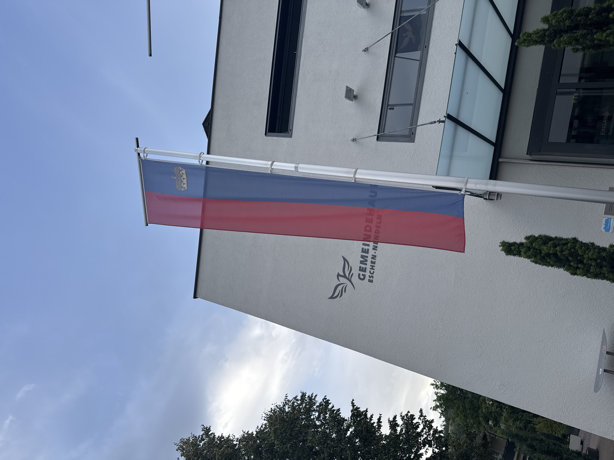 A vertical flag with red on the left and blue on the right, featuring a golden crown, is affixed to a flagpole outside a modern, white building. The building has a sloping facade with a dark grey logo and text reading "GEMEINDEHAUS ESCHEN-NENDELN" which translates to "Municipal House Eschen-Nendeln". The building also features multiple dark-framed windows and a glass awning over an entrance. Evergreen trees are visible in front of the building and to the left side, with a larger tree on the far left. The sky is overcast with scattered clouds. The scene depicts the municipal building in Nendeln, Liechtenstein.