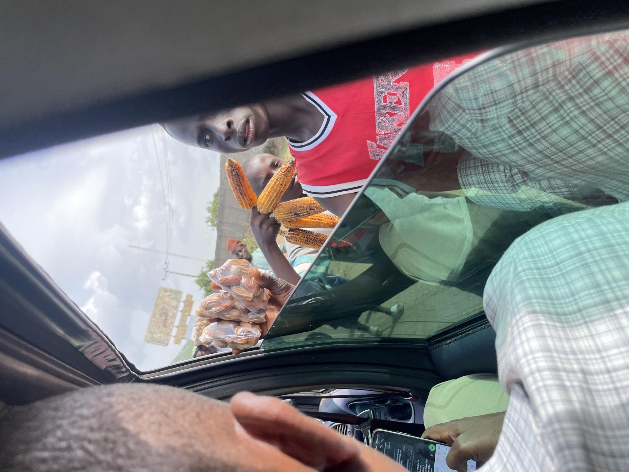 The image shows a group of people standing outside a car with its sunroof open, likely on a road in Jos, Nigeria. The perspective is from inside the car, looking up and out.
The main subject in the foreground, viewed from the chest up, is a young man wearing a red and white basketball-style jersey. He is holding several cobs of roasted corn. Behind him, another person is visible, also appearing to be a vendor. To the right of the main subject, there are packages of what appear to be dried food items, possibly dates or nuts, wrapped in plastic.
The scene appears to be set outdoors during the daytime, under a cloudy sky, suggesting it might be a humid or overcast day. The presence of vendors at the roadside is a common sight in many parts of Nigeria, often catering to passing motorists. The expressions on the faces of the vendors suggest a transactional interaction, with the young man holding the corn looking directly at the camera, perhaps trying to make a sale.
There is some visible text on a phone screen being held by someone inside the car, but it is too small and blurry to be legible. The overall impression is a candid snapshot of a common roadside vending scenario in Nigeria.