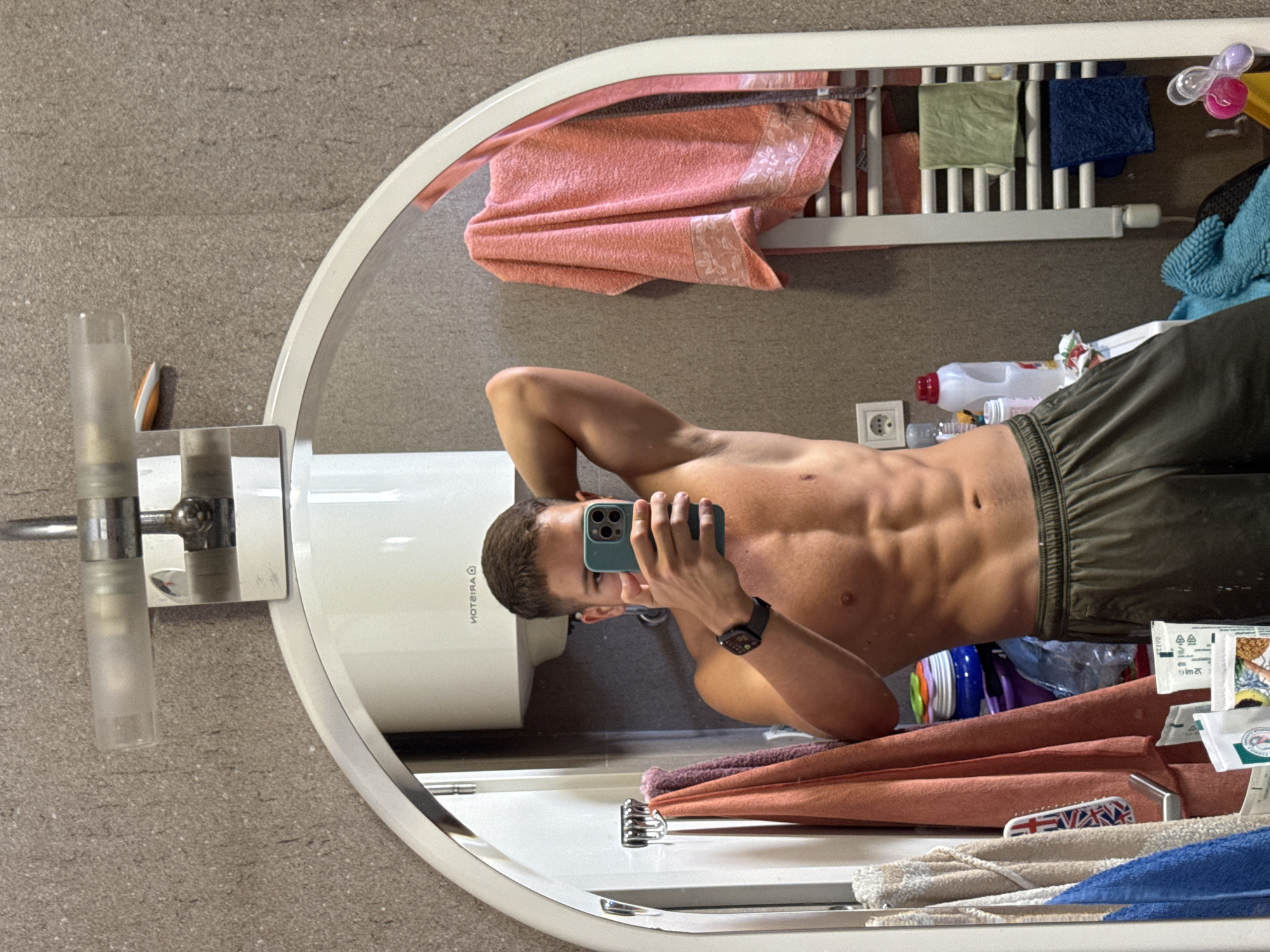 In a bathroom, a shirtless young man with well-defined abdominal muscles and short brown hair is taking a mirror selfie. He is wearing dark green athletic shorts and a smartwatch on his left wrist. His right arm is raised behind his head, flexing his bicep. The reflection in the mirror shows a water heater on the wall and various items on shelves and a towel rack, including towels of different colors, cleaning supplies, and what appears to be a hairbrush. The bathroom has tiled walls. The image was captured in Budva, Montenegro.