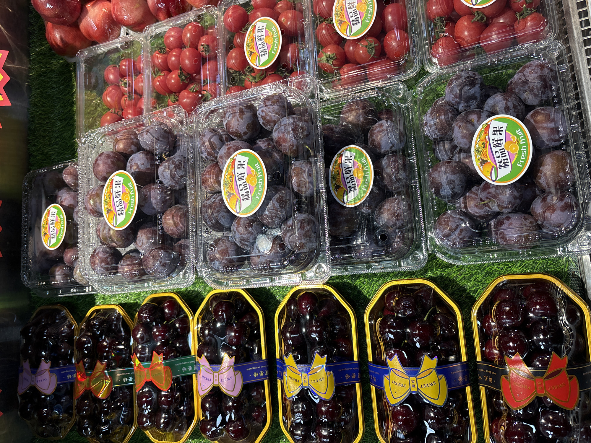 The image displays a close-up view of a retail display filled with various fruits, predominantly cherries and plums, packaged in clear plastic containers. The fruits are arranged on a green artificial grass surface, suggesting a supermarket or fruit stand setting. Several containers feature decorative ribbons and labels, some with Chinese text and the words "Fresh fruit." To the right, a few red fruits, possibly apples or pomegranates, are also visible in a container. The lighting suggests an indoor environment. The scene is devoid of people. The location is identified as Minhang District, China.