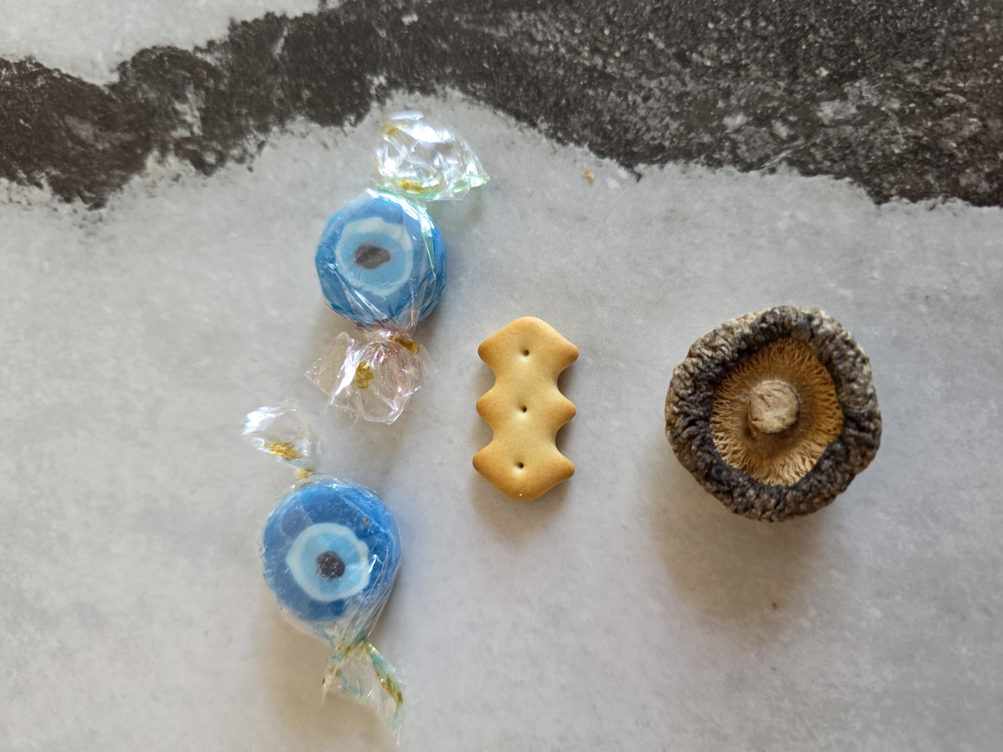 Two blue candies wrapped in cellophane with gold twists on the ends are positioned near the top of the frame. Below the candies, a three-lobed cracker is centered. At the bottom of the frame, a dried mushroom is placed. The objects are on a light-colored, textured surface that appears to be a countertop or table. A dark, irregular shadow or stain runs along the right side of the frame. The scene is indoors, with no view of the outside environment or any indication of the city of Baku, Azerbaijan. There are no people, actions, or interactions depicted.