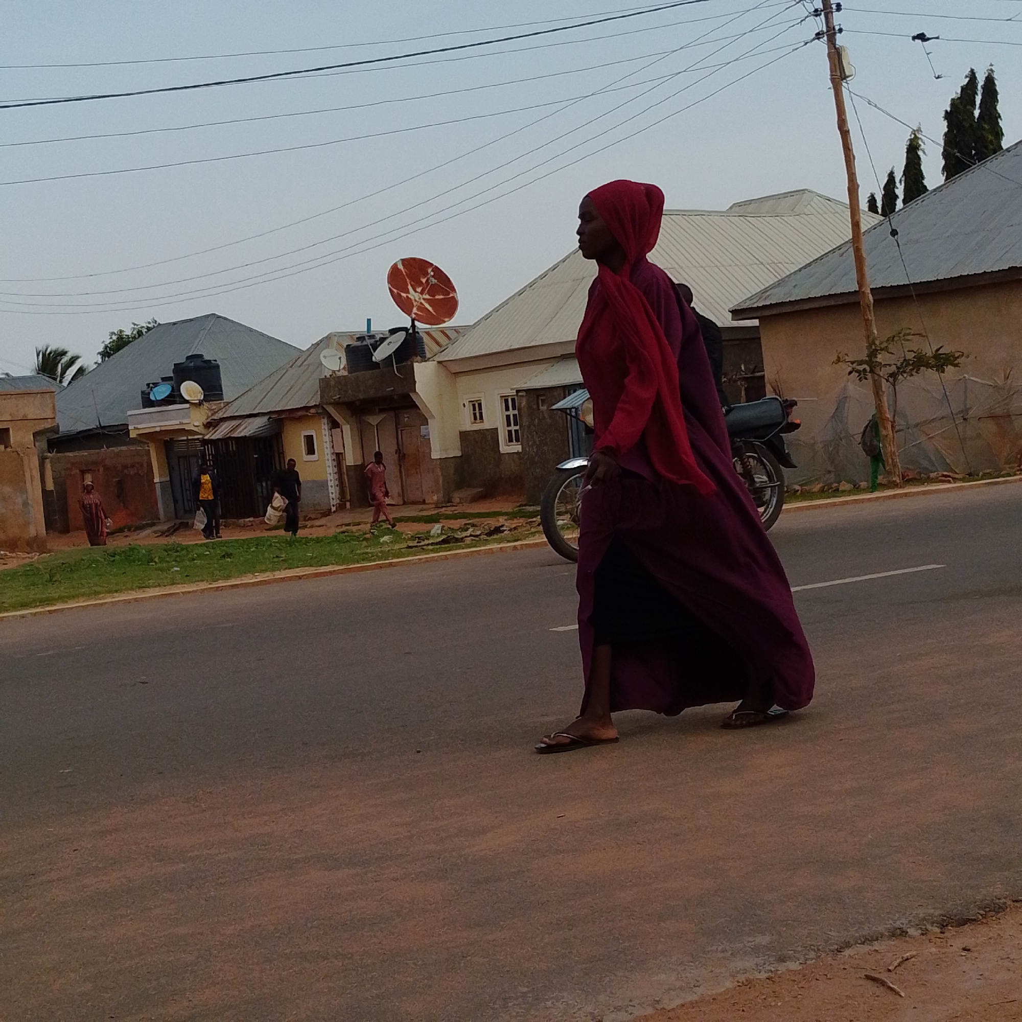 The image depicts a street scene in Gombe, Nigeria. The primary subject is a woman walking across the road from right to left, wearing a vibrant red hijab and a flowing deep purple garment. She is wearing flip-flops. Behind her, to the left, is a motorcycle.
In the background, several residential buildings are visible. These buildings have corrugated metal roofs, common in the region, and some have satellite dishes installed. The houses appear to be constructed with concrete or mud brick. Several other individuals are present in the background, walking or standing near the houses. One person in a yellow shirt is walking towards the right, carrying a white bag. Another person in a pink outfit is walking towards the left, also carrying a bag. A woman in a patterned dress is standing near a building on the far left.
The scene suggests it is late afternoon or early evening, indicated by the soft, diffused light and the shadows. The sky is overcast, suggesting a possibility of rain or generally cloudy weather. The road is paved, with a white line marking the lane. The ground beside the road is a mix of dry earth and some patches of green grass. There is a wooden utility pole with wires stretching across the frame, a common sight in urban and semi-urban areas. No visible text is present in the image. The overall atmosphere is that of daily life in a Nigerian town.
