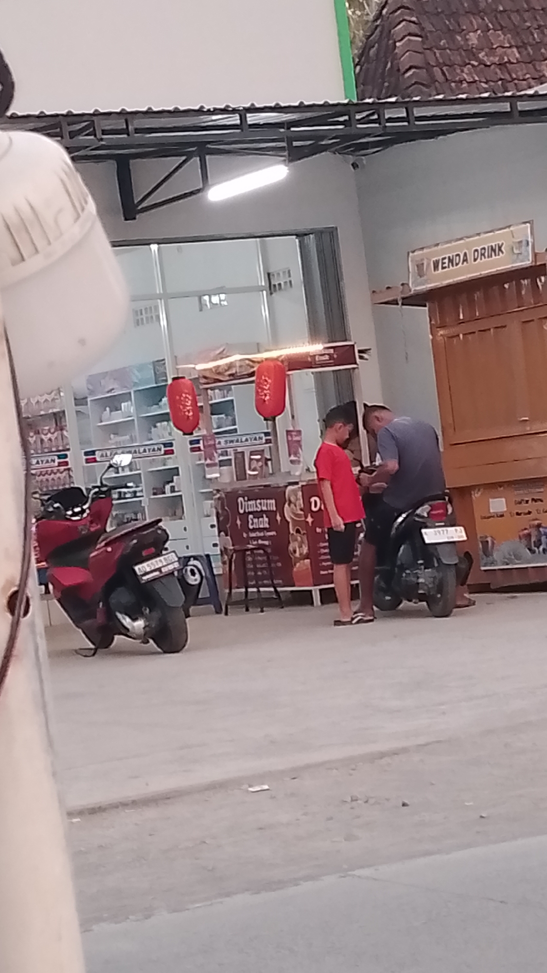 The image depicts an outdoor scene in front of a small shop. A red scooter is parked to the left, and a person is seated on a black scooter to the right, with another person standing next to them. The shop appears to be selling food and drinks, with a sign that reads "Dimsum Enak" (Delicious Dimsum) and "WENDA DRINK." The background shows shelves stocked with various items, typical of a convenience store. The overall setting suggests a casual street-side establishment, possibly in Karanglangu, Indonesia, given the context.