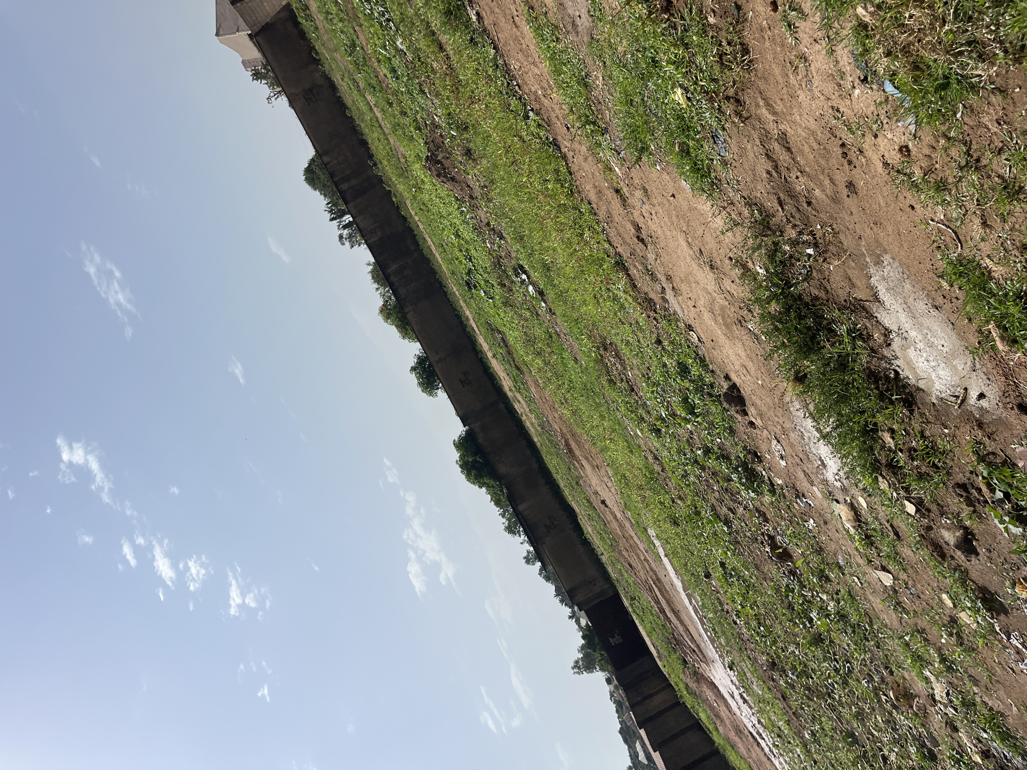 The image is a low-angle, slightly tilted shot of an outdoor scene in Bukuru, Nigeria, under a light blue sky with scattered white clouds. The foreground consists of rough, brown dirt with patches of green grass and some scattered debris, suggesting an undeveloped or neglected area. A faint, wet path or puddle is visible in the lower left portion of the frame. In the mid-ground, the terrain slopes upwards and is covered with more dense green vegetation and some exposed dirt. Dominating the background is a long, grey concrete wall, which appears to be a boundary. Behind the wall, glimpses of trees and some buildings with terracotta-colored roofs are visible. The scene suggests a semi-urban or peri-urban environment, possibly on the outskirts of the city. There are no people or animals visible in the image.