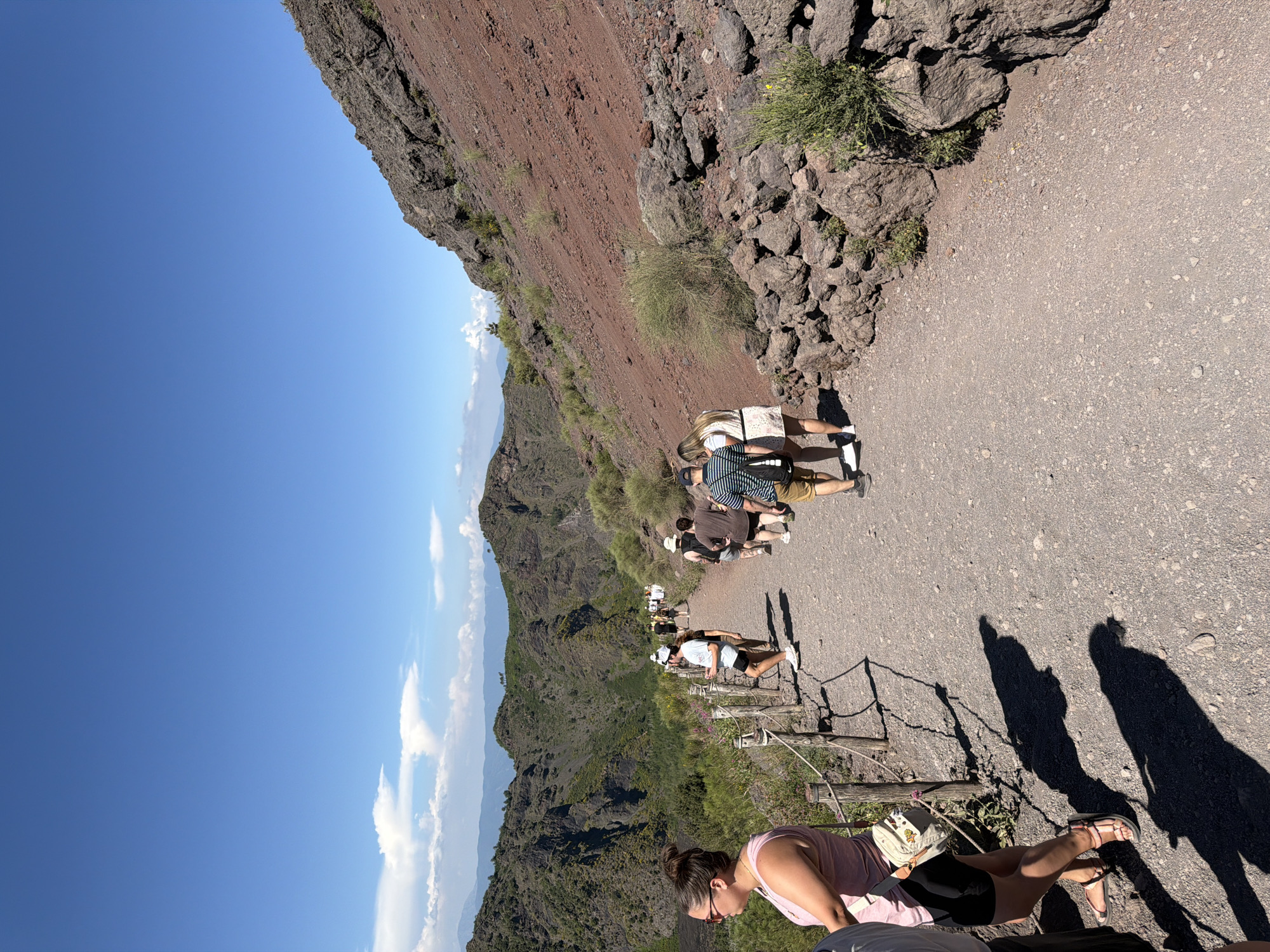 The image shows a group of tourists walking on a gravel path along the rim of a volcanic crater in Ercolano, Italy. The path is bordered by a wooden fence on the left and a steep, rocky slope on the right. The slope is covered in reddish-brown volcanic soil and scattered rocks. In the background, lush green mountains and valleys can be seen under a clear blue sky with scattered white clouds. The tourists are casually dressed for a warm day, with some wearing hats and sunglasses. They are walking in the same direction, seemingly on a guided tour. The overall scene depicts a popular tourist attraction with a natural volcanic landscape.