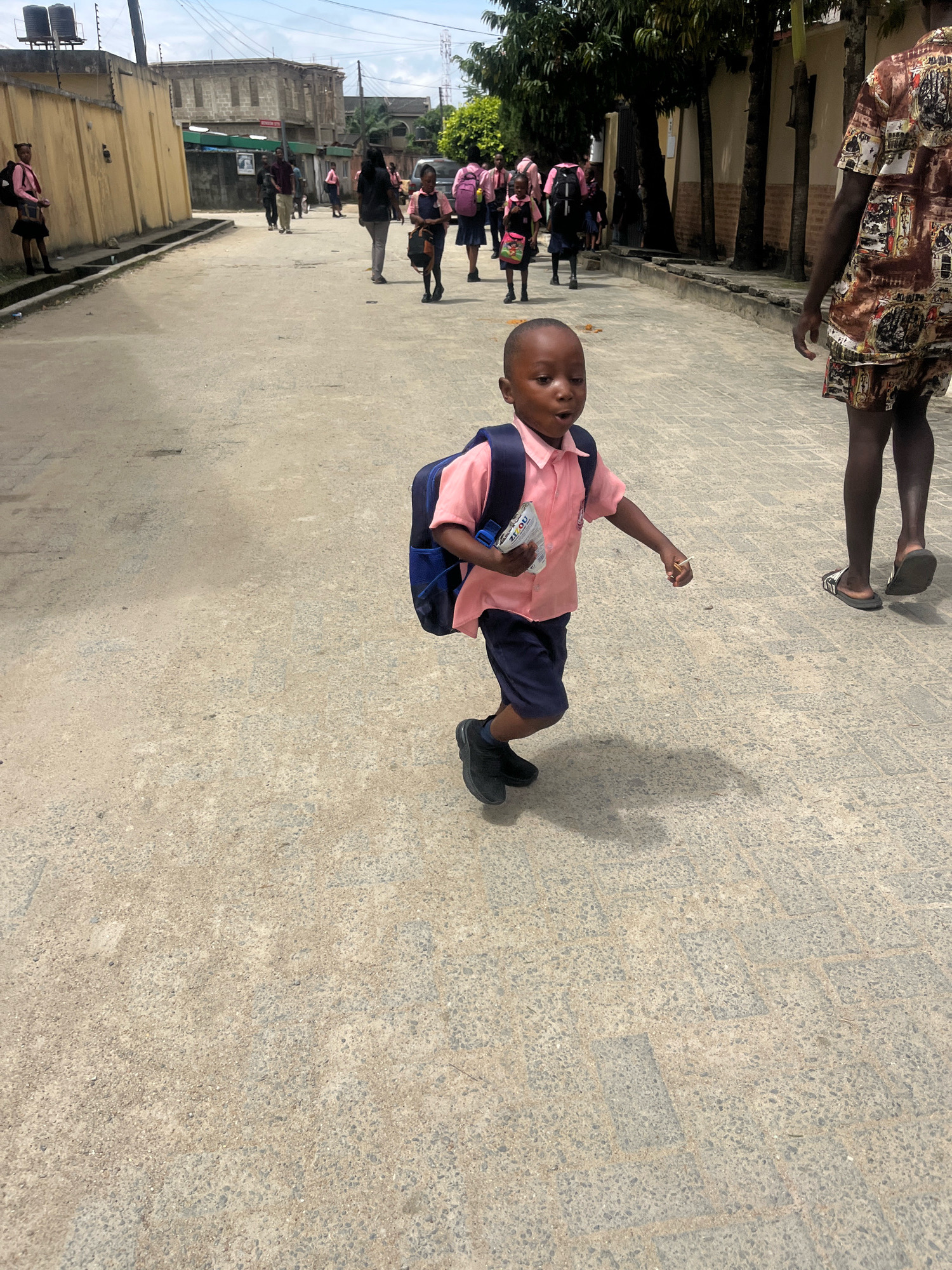 A young boy wearing a pink collared shirt, navy blue shorts, and black shoes, with a navy blue backpack, runs across a paved street in Ado, Nigeria. He holds a white object in his right hand. In the background, other children in similar school uniforms walk along the street. A person in a patterned shirt and shorts walks to the right of the boy. Buildings and trees line the street, and the sky is overcast.