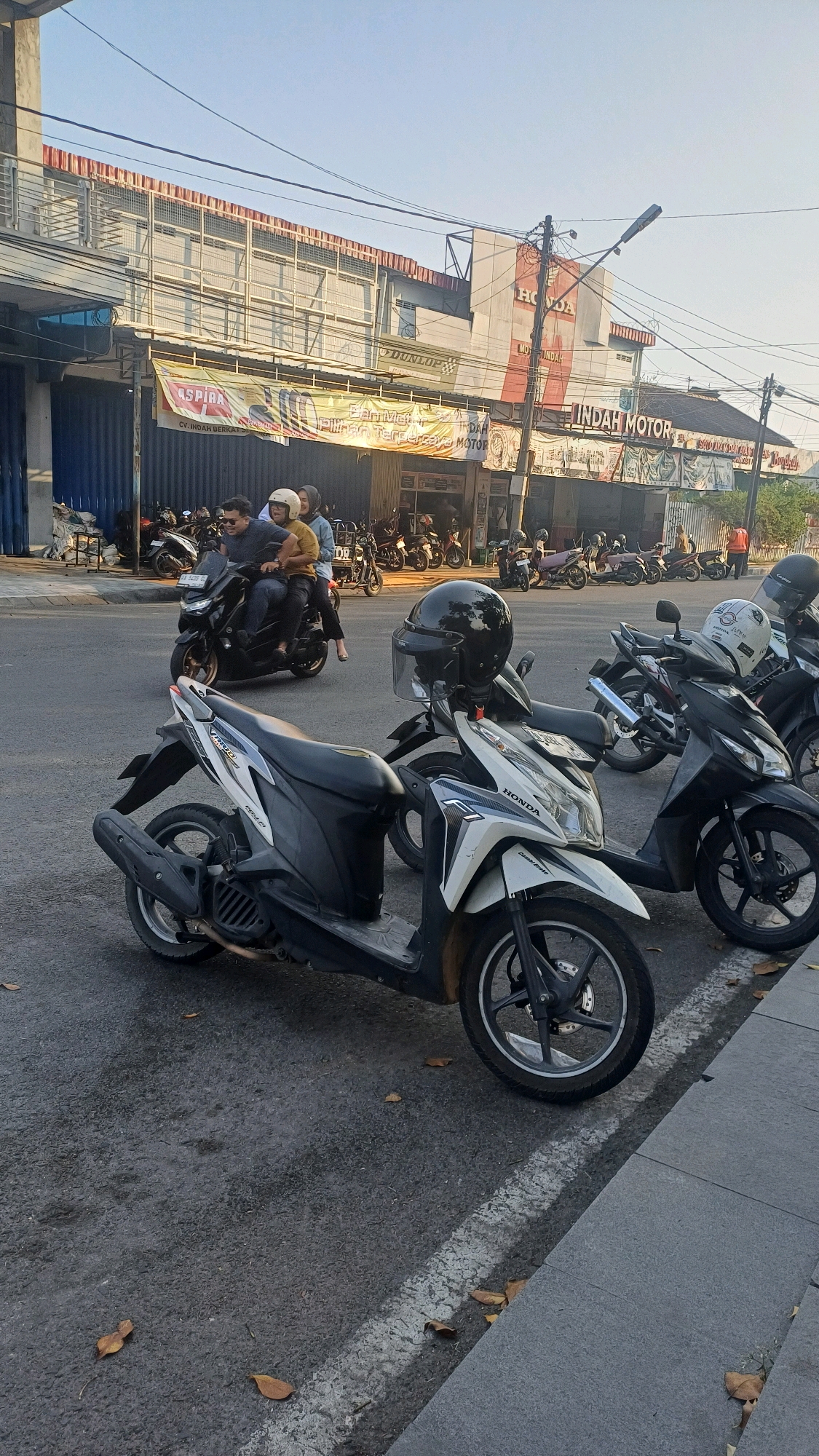 The image depicts a street scene in Klaten, Indonesia, with a focus on parked scooters and a few people. In the foreground, a white and black scooter with a black helmet on its handlebars is parked on the asphalt road. To its right, a black scooter is also parked, with a white helmet on its handlebars. Further back, a black scooter carries three passengers: a man in the front, a woman in the middle, and another person behind her. The man is wearing sunglasses, and the woman in the middle is wearing a yellow shirt and a white helmet. The background shows a row of buildings, including a "HONDA" dealership and other businesses with banners. The sky is clear and blue, suggesting a daytime scene. Several other scooters are parked along the street in front of the businesses. A white line is painted on the road, separating the parking area from the traffic lane. Fallen leaves are scattered on the road and sidewalk.