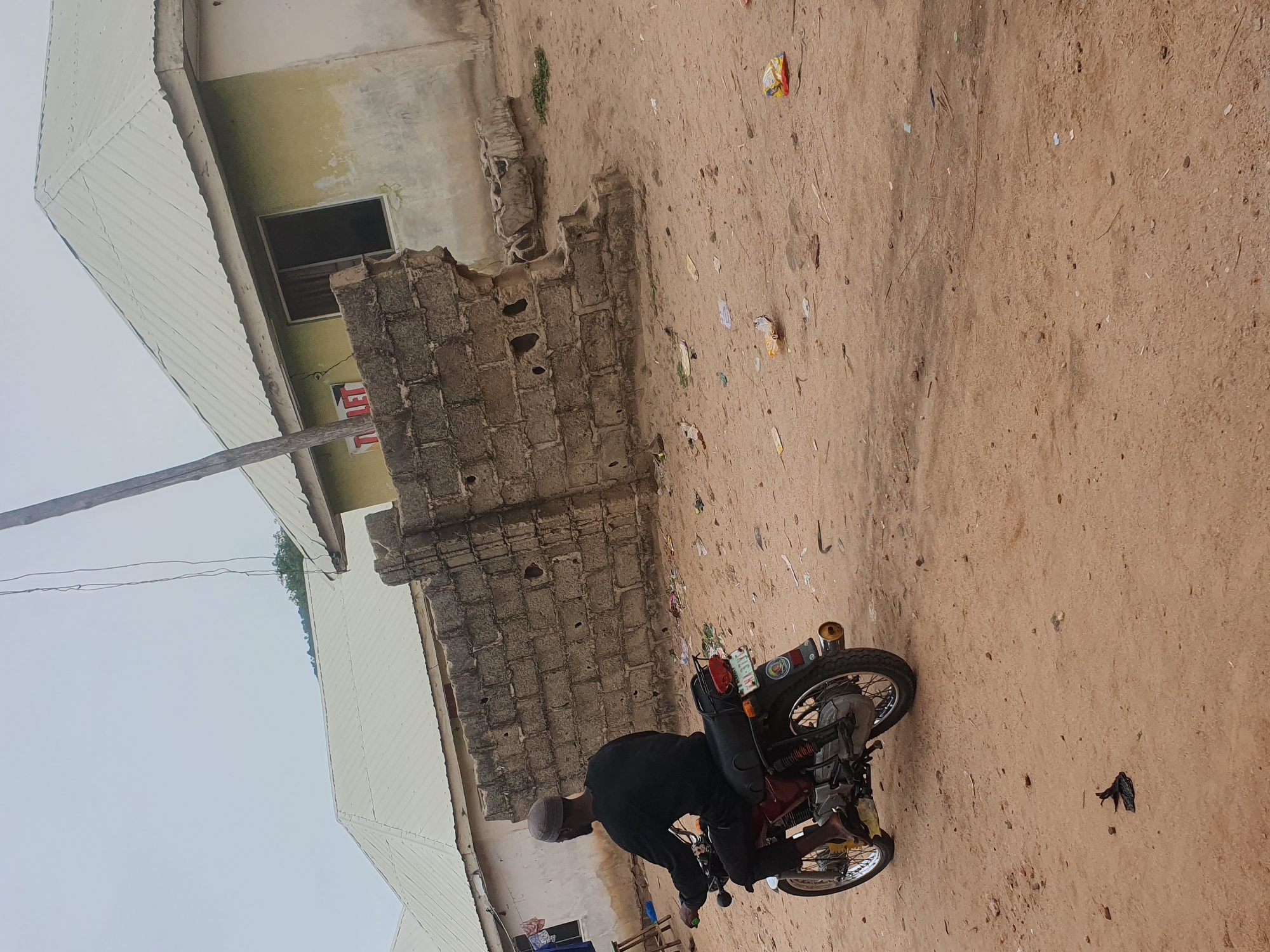 A ground-level, tilted, medium-shot captures a man on a red motorcycle in what appears to be a street in Lokoja, Nigeria. The street is unpaved and covered in a light-brown dirt or sand. Scattered debris, including pieces of paper and plastic, are visible on the ground. The man is dressed in dark clothing, with a visible light-grey or beige cap on his head. He appears to be bending over or working on the motorcycle. The motorcycle has a license plate and other attachments on its rear. To the left, buildings with dilapidated exteriors are visible. The buildings have a mix of brick and painted surfaces, with visible signs of weathering and wear. Some of the structures appear unfinished or partially collapsed. The sky is overcast, creating a muted or diffused lighting across the scene. A pole is visible on the left side of the image.