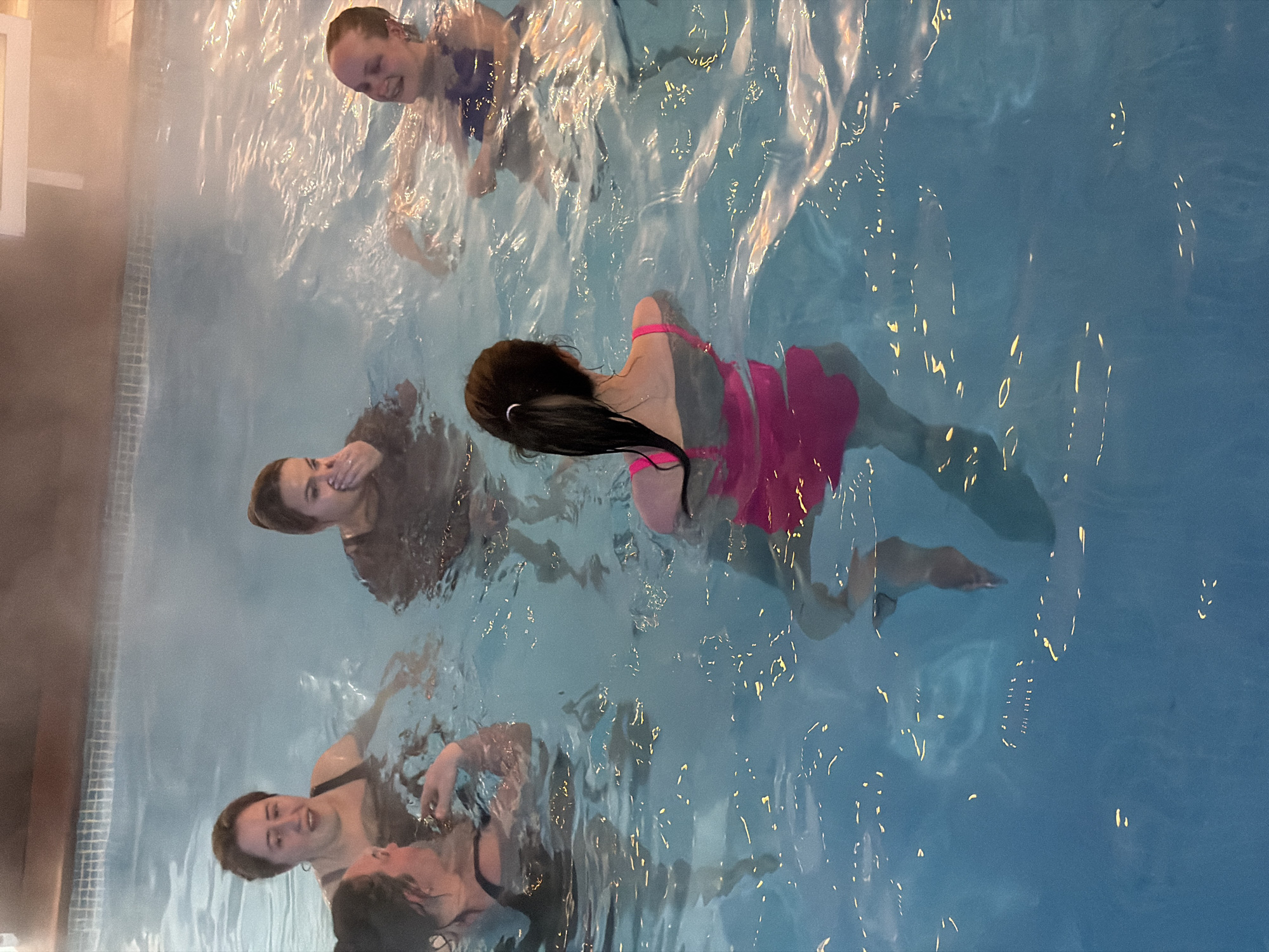 This image captures five young women enjoying an indoor swimming pool in Chaikovsky, Russia. The setting appears to be a warm pool or spa, as indicated by visible steam rising from the water, particularly along the left side of the frame. The pool water is a clear blue, with numerous reflections of overhead lighting creating shimmering patterns on its surface.
In the upper left of the pool, a woman with light hair is seen smiling, wearing a blue swimsuit or top. To her right, another woman with dark hair is visible from behind, wearing a bright pink swimsuit with a ruffled bottom. Her left arm and leg are partially submerged in the water.
In the lower section of the pool, three more women are present. One woman with light brown hair is smiling and looking upwards, wearing a dark swimsuit. Adjacent to her, another woman with dark hair is also in the water, and her face is visible, seemingly engaging in conversation or interaction with another person whose face is partially visible to her right. All are wearing dark swimsuits.
The overall atmosphere is one of relaxation and enjoyment, with several of the women displaying happy and content expressions. The image captures a leisurely moment among friends in a well-lit indoor aquatic environment, evidenced by the tiled pool edge and wooden paneling visible on the far left. There is no visible text in the image.