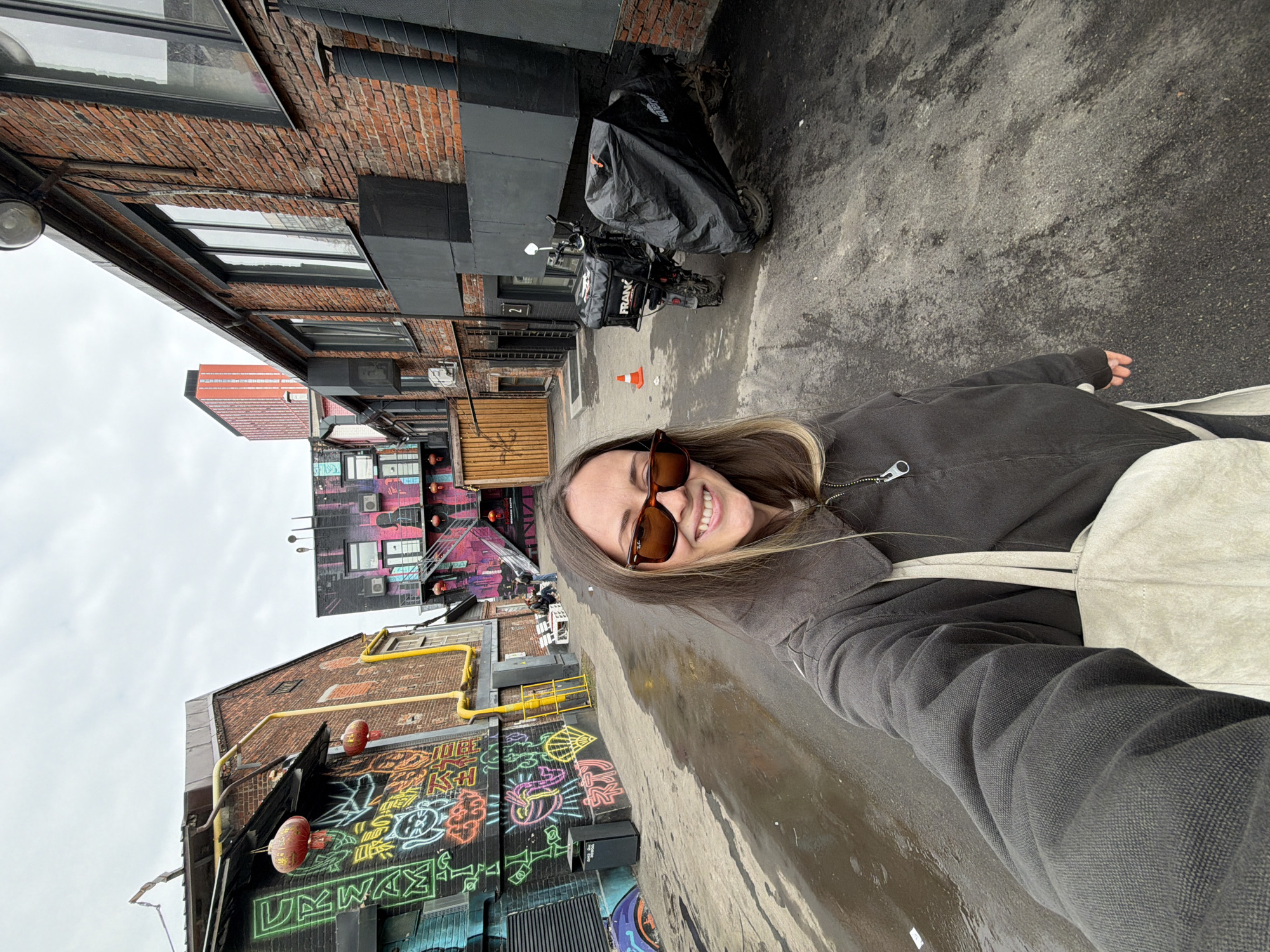 A female subject is depicted in a selfie taken outdoors within an urban alleyway in Khimki, Russia, under an overcast sky. The subject, positioned in the foreground and center, has shoulder-length brown hair with blonde highlights, wears dark sunglasses, a dark grey zip-up jacket, and carries a light-colored tote bag over her left shoulder. She is smiling directly at the camera.
The ground is asphalt, exhibiting various damp patches. To the subject's left, a brick building features extensive, multi-colored graffiti in a neon style, including Asian characters, a cat graphic, and other symbols. Red Chinese lanterns are suspended from an overhang on this building. A black trash bin is situated near the graffiti wall. Further down the alley on the left, three individuals are seated on two white benches.
To the subject's right, brick buildings with large windows are visible, some sections painted dark grey or black. A black delivery scooter or motorcycle is parked against the right wall, with a black "FRANC" branded delivery box on its rear rack. The vehicle's front half is covered by a black "Waterproof" cover. An orange traffic cone stands on the ground adjacent to the scooter.
In the mid-to-far background, additional buildings with colorful graffiti are present, and a prominent red-orange high-rise building is visible in the distance beneath the uniformly grey sky.