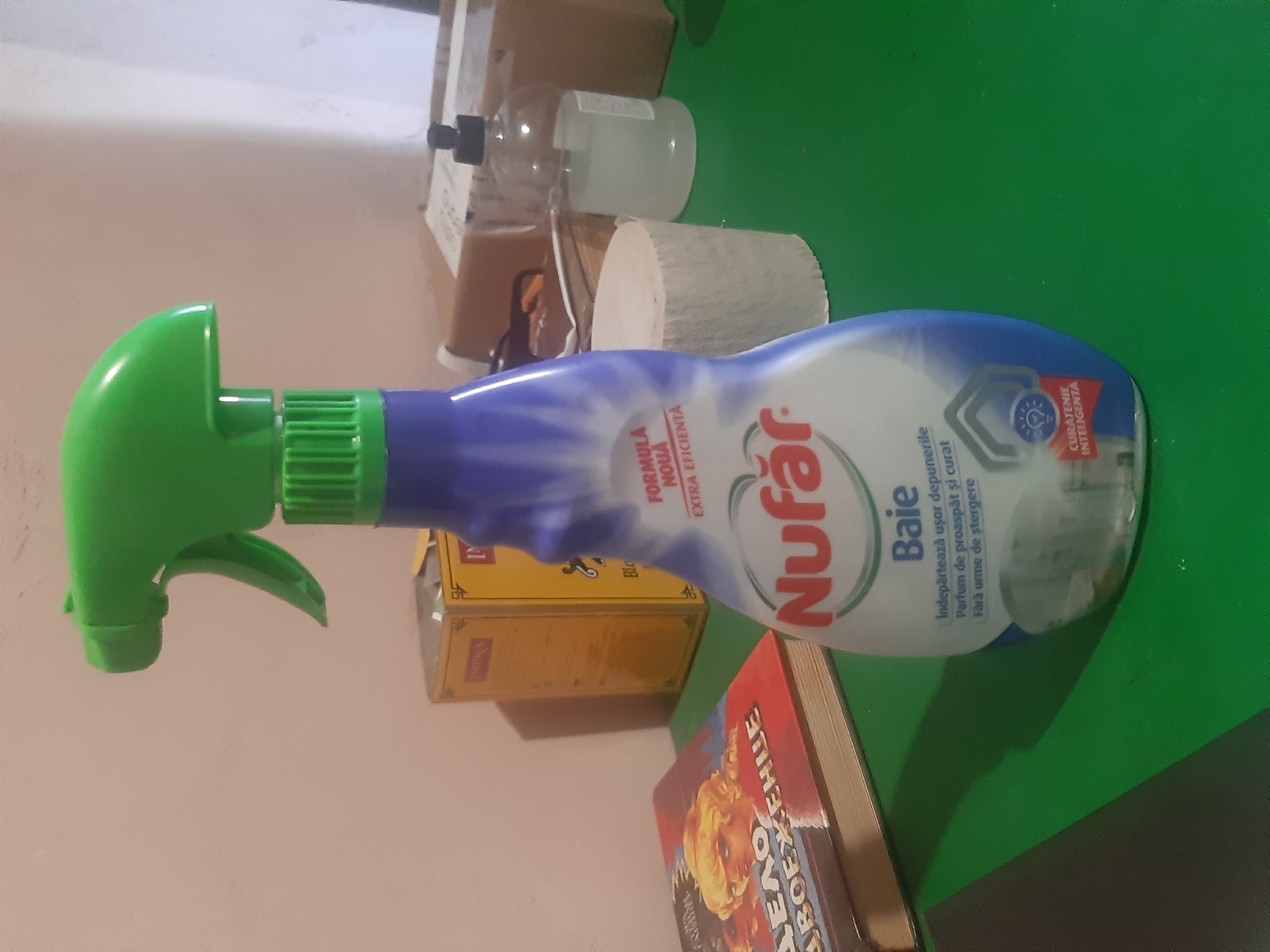 A frontal, medium shot captures a partially filled bottle of Nufăr brand bathroom cleaner, with its signature green trigger spray head and purple-blue bottle, standing on a green surface. The bottle is positioned slightly off-center to the right. Behind it, a roll of toilet paper is visible, along with a clear plastic pump bottle containing a pale liquid and a black pump dispenser. Further in the background, cardboard boxes are stacked. To the left of the cleaner, a closed book with a colorful illustration of a blonde-haired figure is partially visible. The wall behind the objects is a neutral, light color. The overall scene appears to be an indoor setting, likely a home environment, possibly a kitchen or bathroom counter, though the specific location in Cricova, Moldova cannot be determined from the visual information.