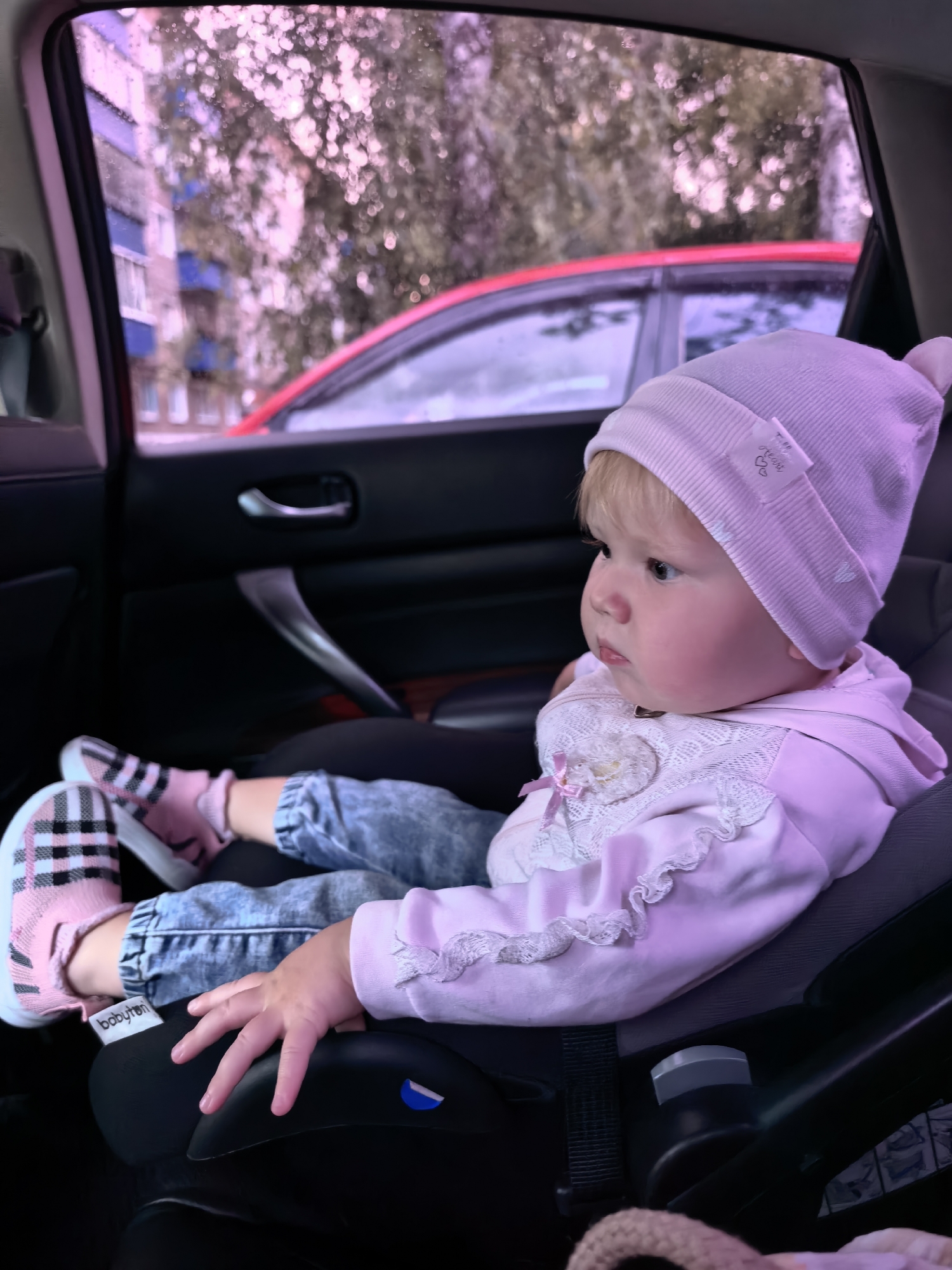 The image shows a young child, likely a toddler, sitting in a car seat within a vehicle. The child is wearing a pink knitted hat and a pink outfit adorned with lace and a small pink bow. The child's feet are clad in pink socks and black and pink plaid slip-on shoes. The child is looking away from the camera, with a focused expression.
The car seat is black and positioned on the backseat of the car. Through the car window, a blurry view of a city street can be seen, with buildings and a red car. The lighting suggests it is daytime. The overall scene is consistent with a child traveling in a car in an urban environment. The crops confirm the presence of a car interior, a child in a car seat, and a view of the outside world through the window, indicating a street scene in the city of Sterlitamak, Russia.