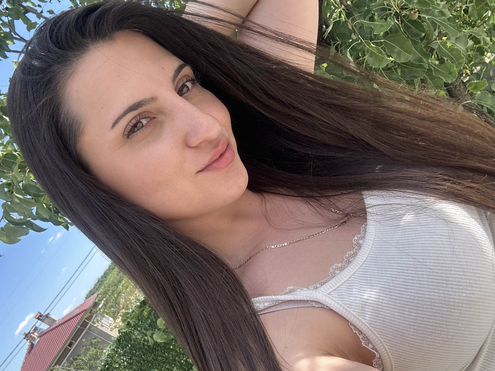 A woman with long, dark brown hair, fair skin, and light brown eyes is posing outdoors in what appears to be a selfie. She is wearing a white ribbed tank top with lace trim and a gold chain necklace. Part of a residential building with a red tile roof and chimney is visible in the background, indicating the location is in a residential area in Beliakovets, Bulgaria, possibly a suburb or outskirts of the town. Green foliage suggests a natural or semi-natural environment. The sky is blue. The woman's right arm is raised above her head.