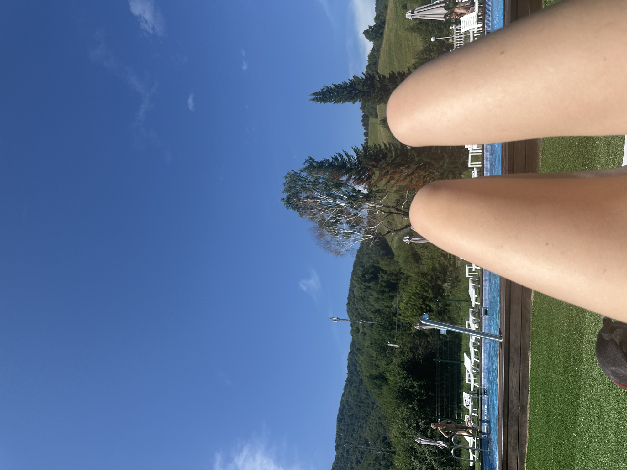 The image depicts a sunny day at an outdoor swimming pool with a mountainous backdrop. In the foreground, a person's legs are visible, suggesting they are lying down and looking towards the pool. The pool area is furnished with white lounge chairs, some unoccupied and others with people relaxing. A woman in a bikini stands near the edge of the pool. Behind the pool, lush green trees cover the rolling hills, and a clear blue sky with scattered white clouds dominates the upper portion of the frame. A tall communications tower is visible on one of the hills. The overall scene suggests a resort or recreational area.