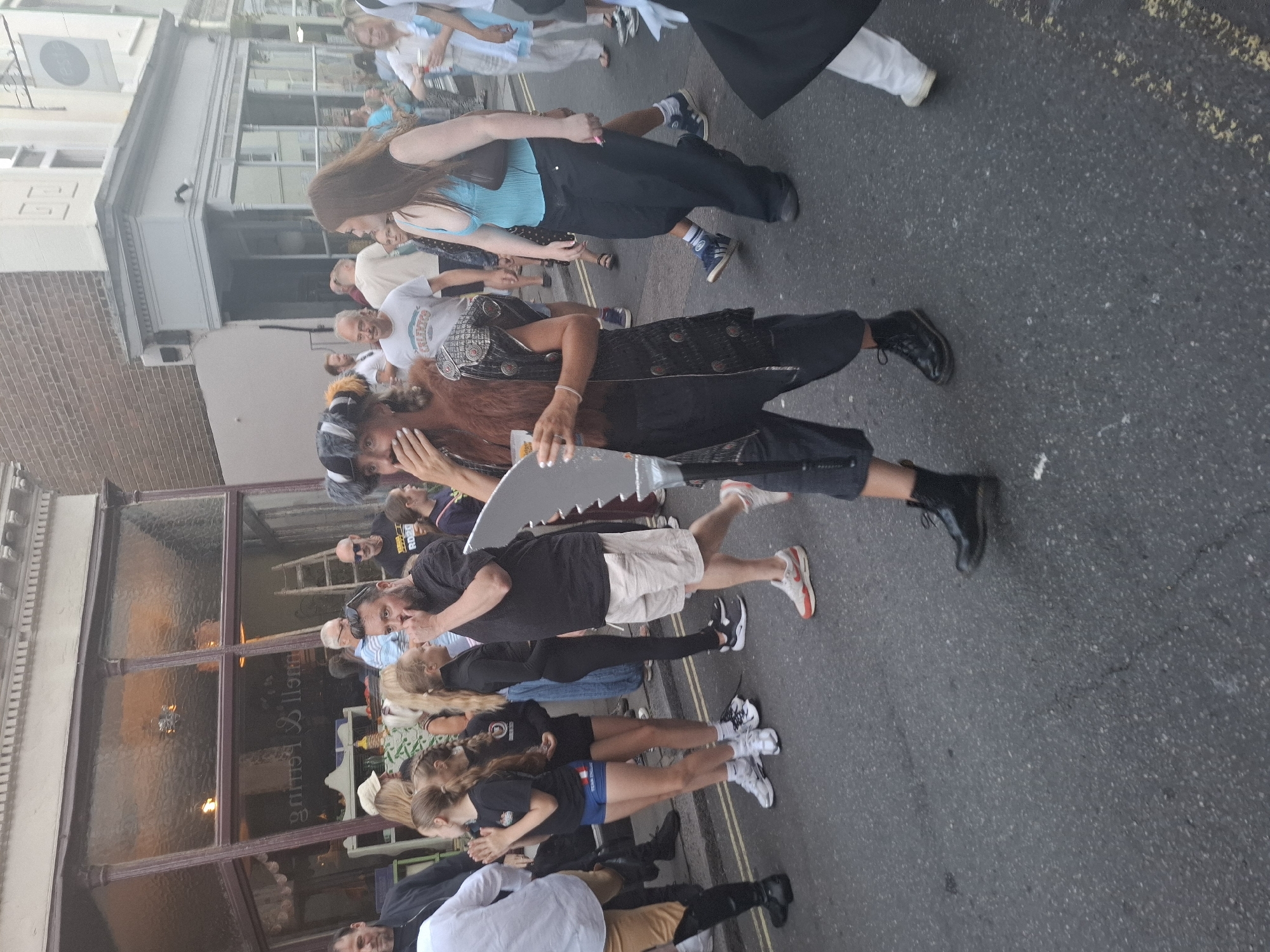 This is a street-level, medium shot taken from a slightly low angle in the city of St Leonards, United Kingdom. The scene depicts a group of people walking on an asphalt street. In the foreground, a person in costume is walking towards the right, holding a drink and a large, metallic, sword-like object. The costume includes a furry collar, a patterned vest, and dark pants. To the left, two young females in athletic wear are walking, with one clapping. Behind them, an adult male in a black t-shirt and shorts is walking, and another adult male in a black jacket is visible on the far left. In the background, several other individuals are walking and standing, some with drinks in hand. Storefronts with large glass windows line the street on the left side of the image. The lighting suggests it is either late afternoon or early evening.