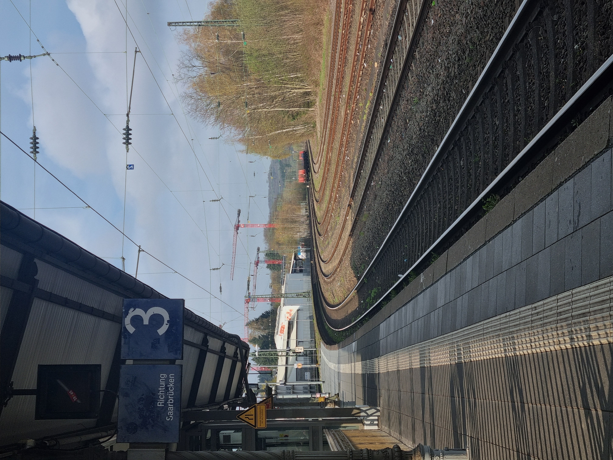 This image depicts a railway platform at a train station in Sankt Ingbert, Germany. Platform 3 is indicated by a sign. The destination "Richtung Saarbrücken" (towards Saarbrücken) is also displayed. Railway tracks curve into the distance, with several tracks visible. The train station includes overhead electrical lines and support structures. A forest area is visible along the tracks. The station platform is paved with gray tiles. Red construction cranes are present in the middle distance.