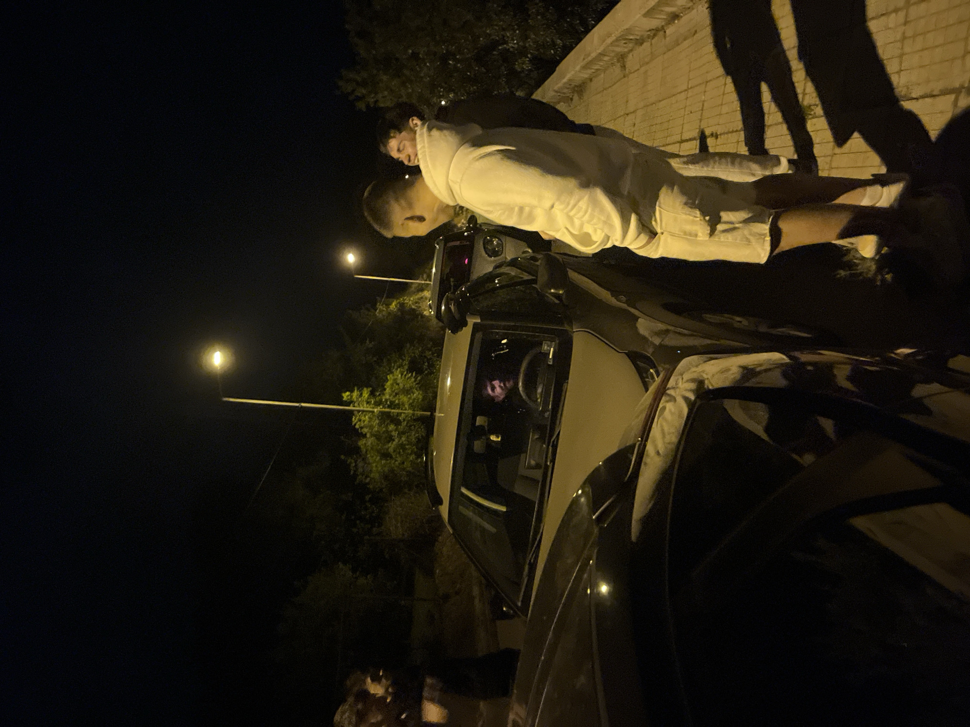 At night in an urban area of Unknown, Unknown, a person in a white outfit is leaning over the hood of a dark-colored car. Another person is standing next to them. A street light is in the background, illuminating the area and some trees. The shadows of people are visible on a light-colored wall behind the individuals.